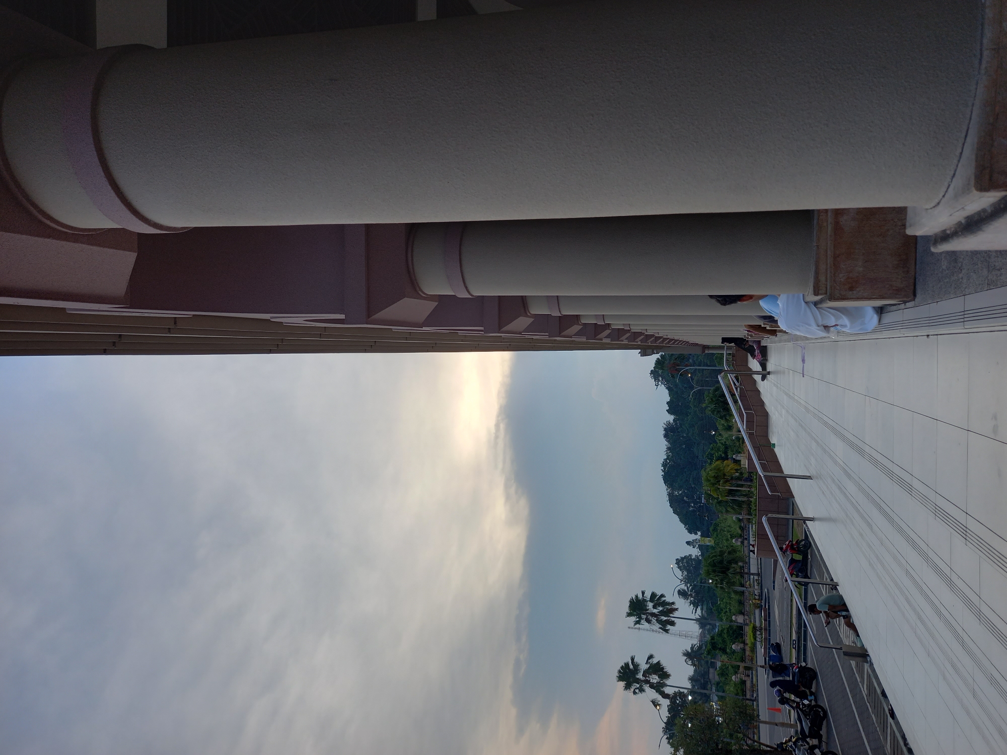 This photograph captures an exterior view of a building with a colonnade of large, cylindrical columns, likely a mosque or a public institution, in Samarinda, Indonesia. The columns are light-colored and textured, with subtle pink banding near the top. The building's facade appears to be a muted pink or mauve. In the foreground, a wide, tiled walkway with a subtle pattern slopes upwards and to the left. A metal railing runs along the edge of the walkway.
Several people are visible, primarily seated or lounging on the steps and walkway. One person in the lower right is wearing a white garment. Further left, individuals are seated near the railing. In the middle ground, there's a parking area with several motorcycles, suggesting a public space. A few individuals are interacting with the motorcycles, and others are seen sitting or standing in the distance.
The background features a landscape with palm trees, lush green foliage, and more distant trees on a gentle slope. The sky is overcast with soft clouds, allowing a gentle, diffused light to illuminate the scene, hinting at either dawn or dusk. The overall atmosphere is serene and somewhat subdued, indicative of a quiet moment in a public space in Samarinda.