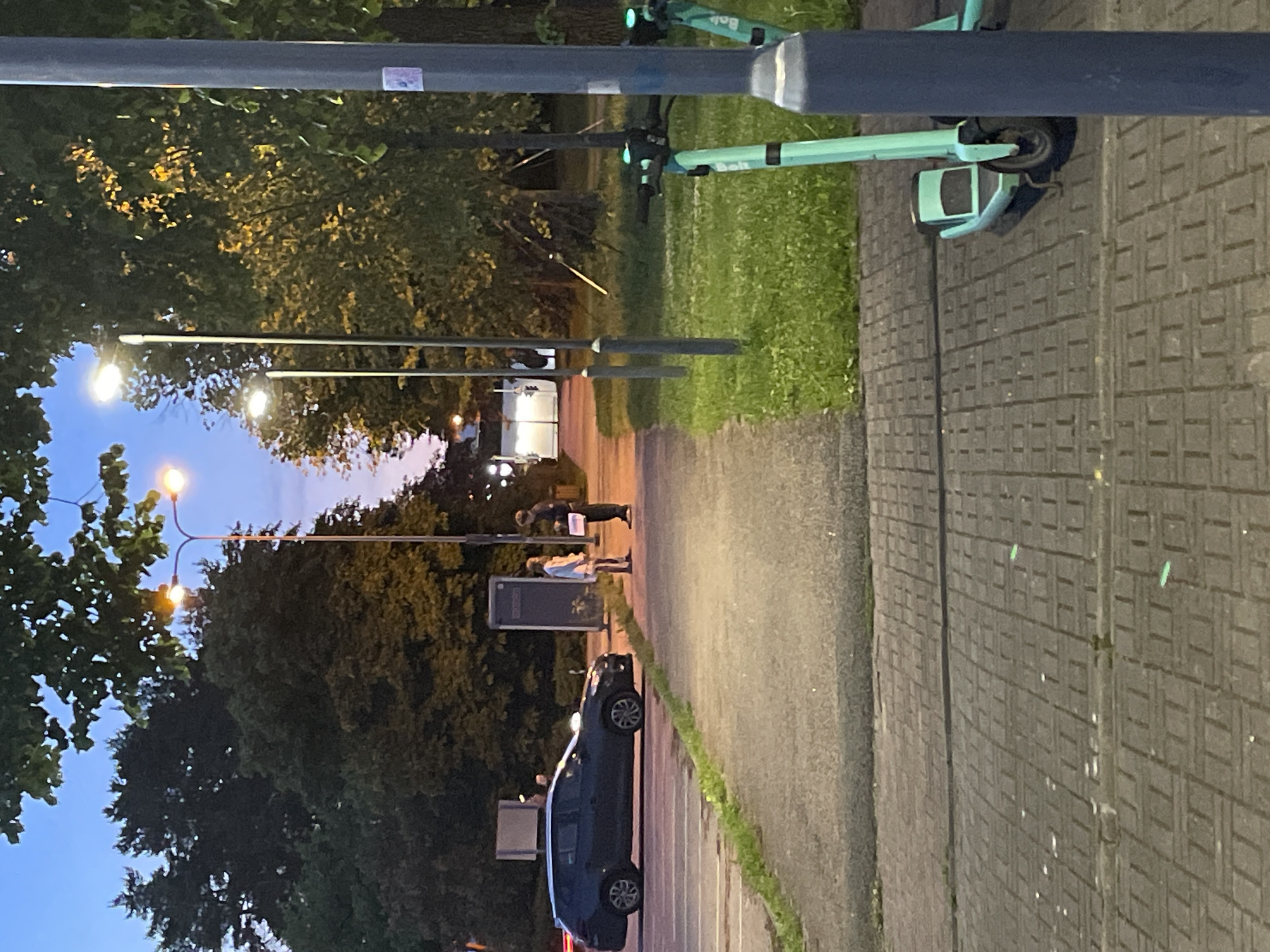 The image shows a street scene in Tallinn, Estonia, at dusk. A dark-colored hatchback car is parked on the left side of the road. On the sidewalk, two people are standing, facing each other. The person on the left is wearing a light-colored outfit, and the person on the right is wearing dark clothing and holding a white object. Several streetlights are visible, some of which are illuminated. In the foreground on the right, two green electric scooters are parked. The environment includes trees, grass, a paved walkway, and a brick-patterned ground surface. The sky is a dusky blue with hints of pink near the horizon.