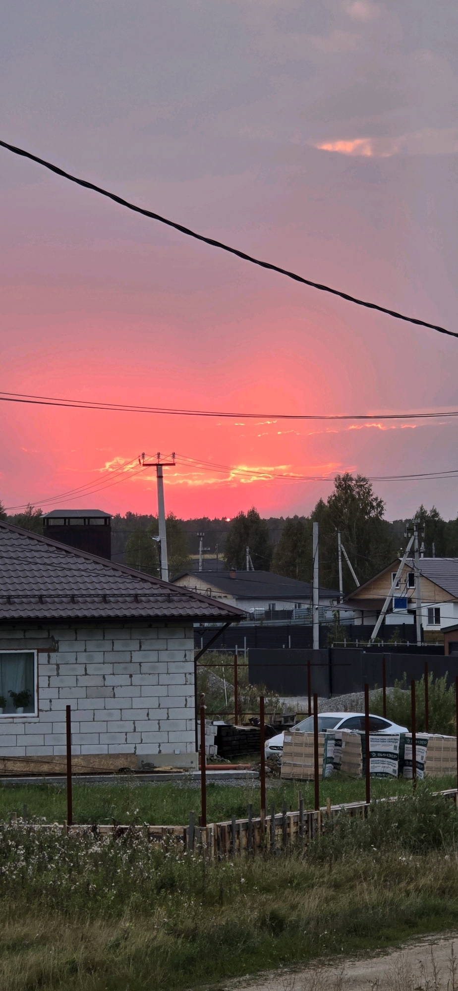 The image shows a rural landscape at sunset. The sky is a vibrant pink and orange, with some clouds. In the foreground, there is a partially constructed house made of white bricks. There are also power lines and utility poles visible. In the background, there are trees and other houses. The scene appears to be in a residential area under construction, possibly in the city of Klyuchi, Russia, given the presence of Cyrillic text on some packaging in the foreground. There are no people visible in the image. The environment suggests an outdoor setting during twilight hours.