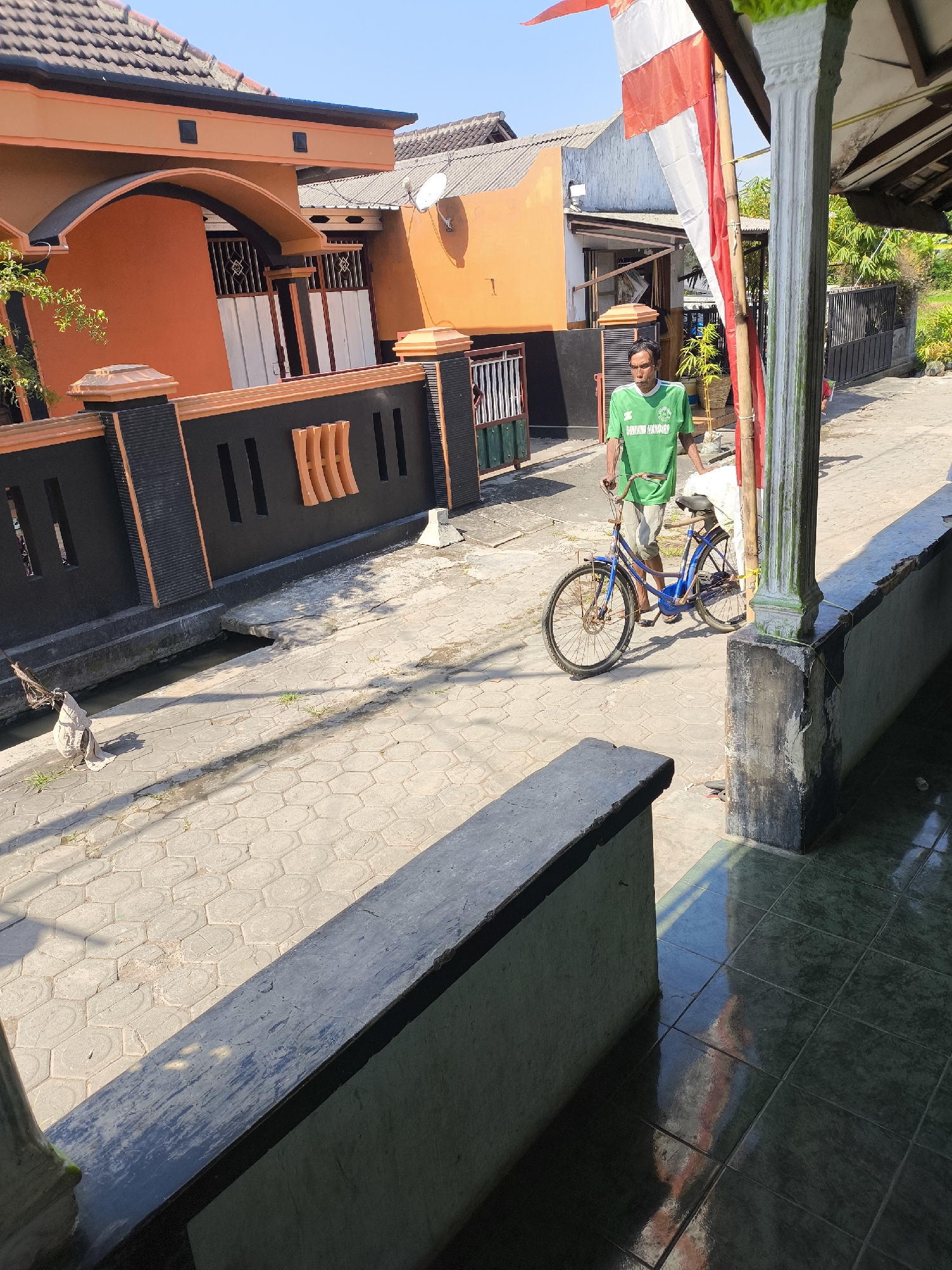 A medium shot captures a street scene in Lumajang, Indonesia, bathed in bright daylight under a clear blue sky. In the foreground, a weathered concrete structure juts into the frame from the bottom left, its surface showing signs of age and cracking. To the right, a tiled veranda with dark green tiles reflects the sunlight, creating a glossy sheen. A thick, grey concrete pillar, weathered and stained, stands prominently on the right side of the image.
Dominating the mid-ground is a man wearing a bright green t-shirt and light-colored shorts. He is standing next to a blue bicycle, with his right hand resting on the handlebars. The bicycle appears to be a simple, utilitarian model with a step-through frame. The man is looking slightly to his left, his expression neutral. Behind him, a narrow street paved with hexagonal concrete blocks stretches into the distance.
To the left of the street, a low black fence with orange accents separates the sidewalk from a shallow, water-filled drainage ditch. Beyond the fence, an orange house with a curved archway and white and grey doors is visible. The house has a tiled roof, and a satellite dish is mounted on an adjacent orange building. A red and white Indonesian flag, attached to a tall pole, is partially visible on the right side of the frame, fluttering gently. The environment suggests a residential area, likely in a tropical climate, given the bright sun and lush greenery hinted at in the background. The overall impression is one of everyday life unfolding on a typical day in Lumajang.