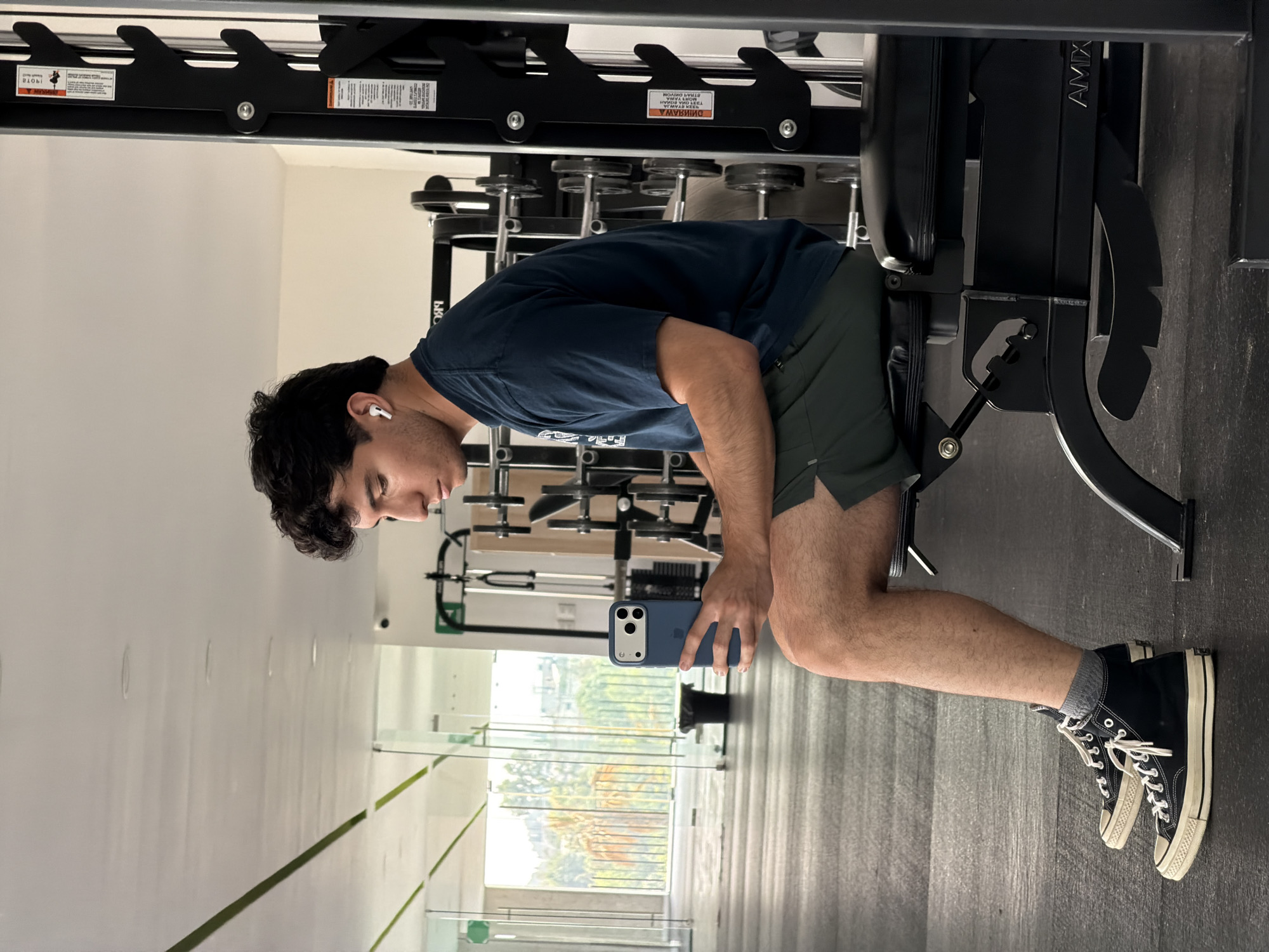 A young adult male is in a gym setting. He is bent over, looking at a blue-cased smartphone he holds in his hands. He has dark hair, wears an earbud in his left ear, a dark blue t-shirt, and dark green shorts. He has on dark high-top sneakers with white laces and gray socks. He is positioned near a piece of gym equipment. In the background are rows of weights and other exercise machines. Outside of a wall of glass, it's possible to see trees. The city of Santa Clara Ocoyucan, Mexico, cannot be determined from the image.