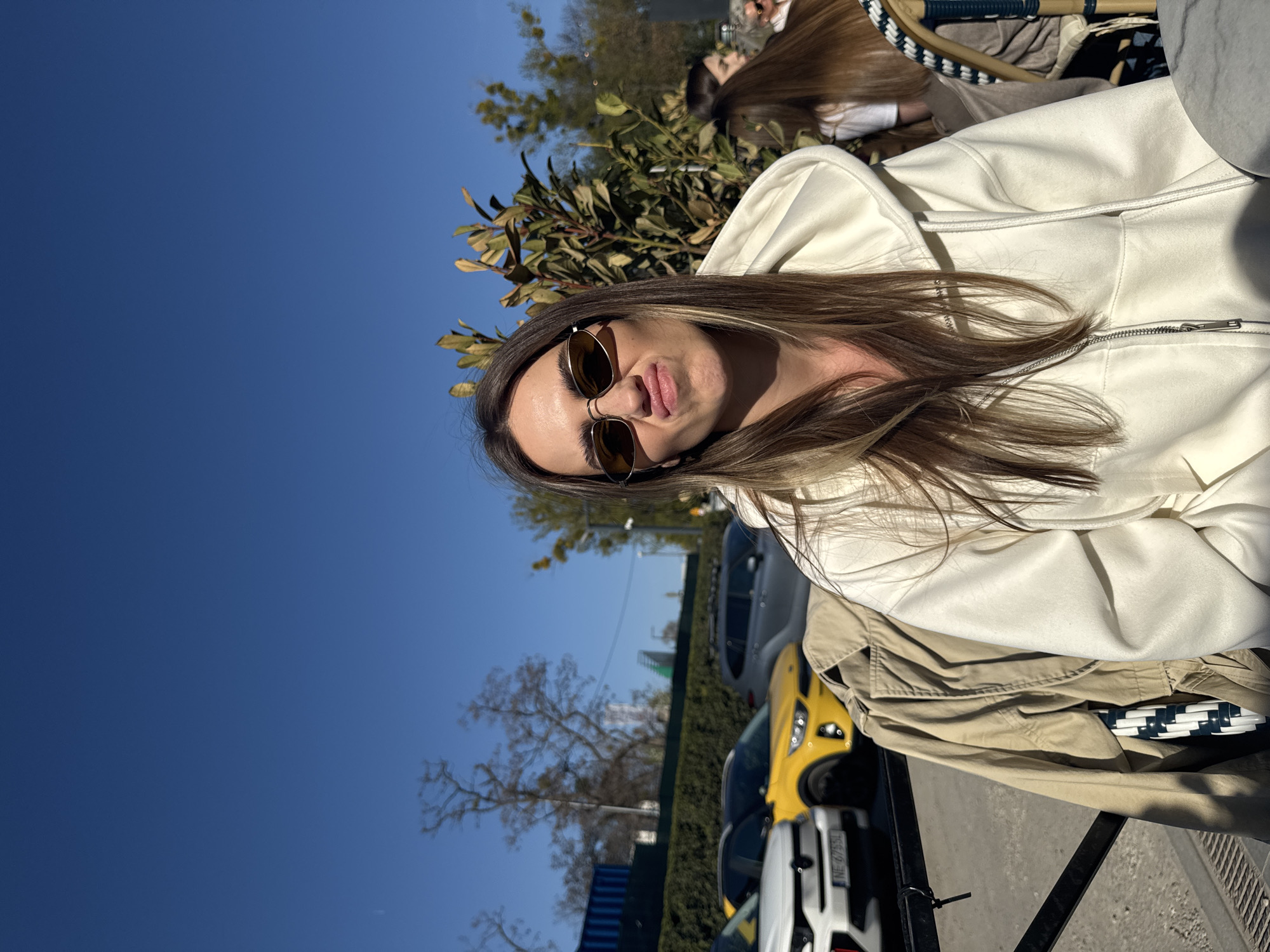 The image depicts a woman seated outdoors at what appears to be a cafe or restaurant. She wears sunglasses and a white zippered hoodie. Her hair is long and brown with lighter highlights. She is making a pouting facial expression. In the background, there are parked cars, including a yellow one, and trees against a clear blue sky. Another person is partially visible in the background. Based on available information ( license plates visible in image) the location is in Warsaw, Poland.