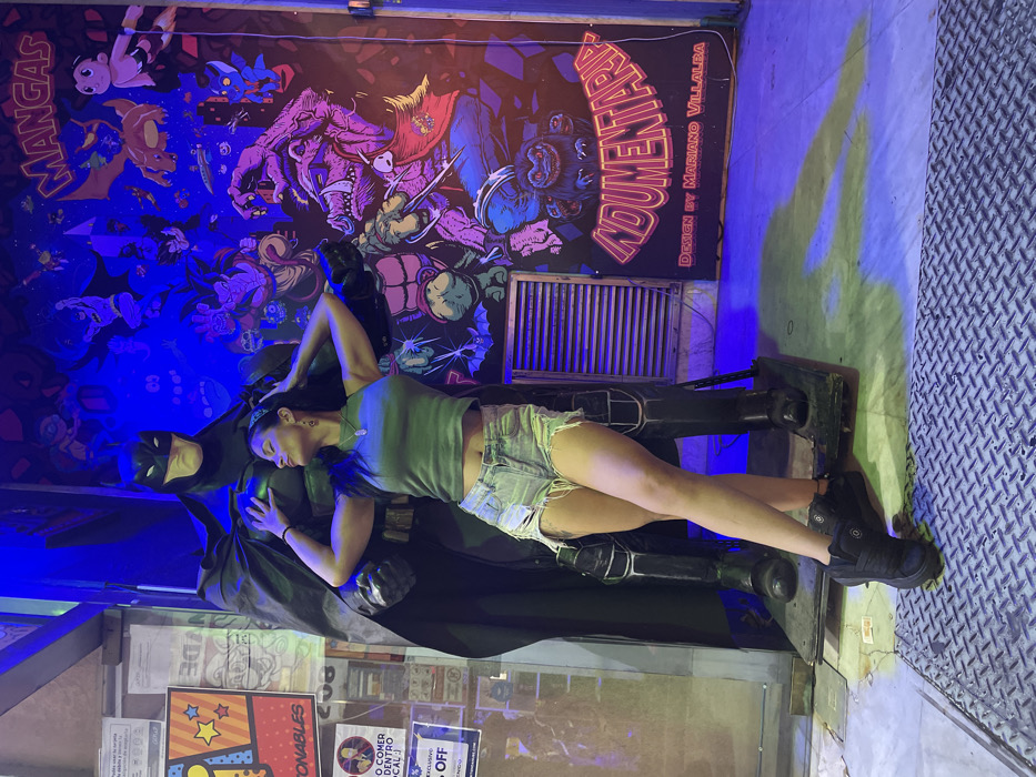 A woman in a green top and denim shorts leans against a Batman statue. A wall mural with comic book-style illustrations in vibrant blues and purples is in the background. Signage is affixed to the wall under the mural, and the scene is lit with a blue light. The floor has a diamond plate pattern.
