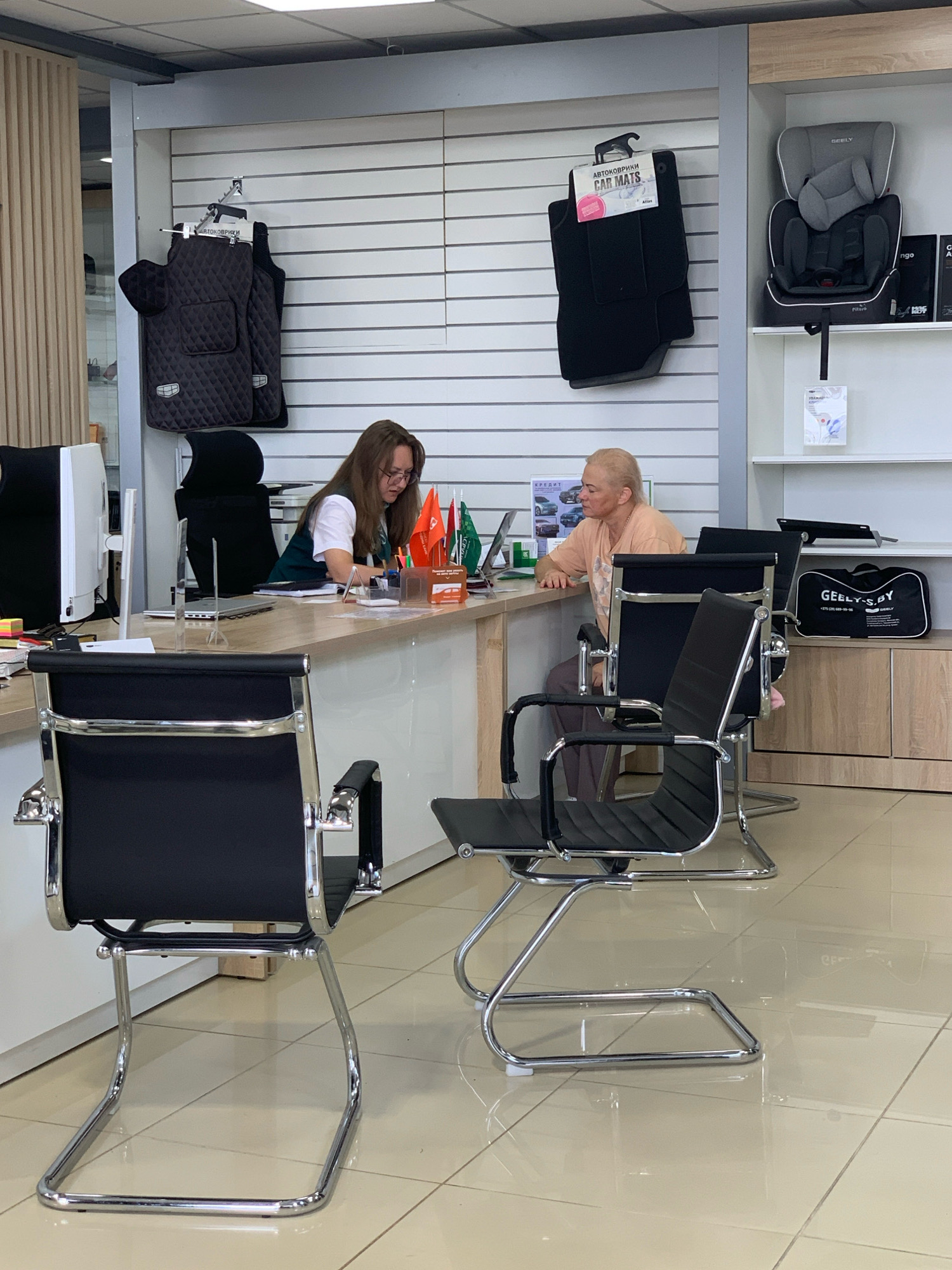 A retail showroom interior, possibly a car dealership, is depicted. Two people are present: a woman in a white shirt and dark vest is seated behind a counter, looking at a laptop, and a person with a shaved head, wearing a light orange t-shirt, is seated on the opposite side of the counter, facing the woman and also looking at a laptop. The environment includes a counter with office supplies, flags, and promotional materials. Shelving units display car accessories like car mats and a child car seat. The floor is tiled, and office chairs are positioned in the foreground. The overall impression is of a commercial space dedicated to automotive sales or related services. The location is identified as Чыжэвіцкі сельскі Савет, Belarus.