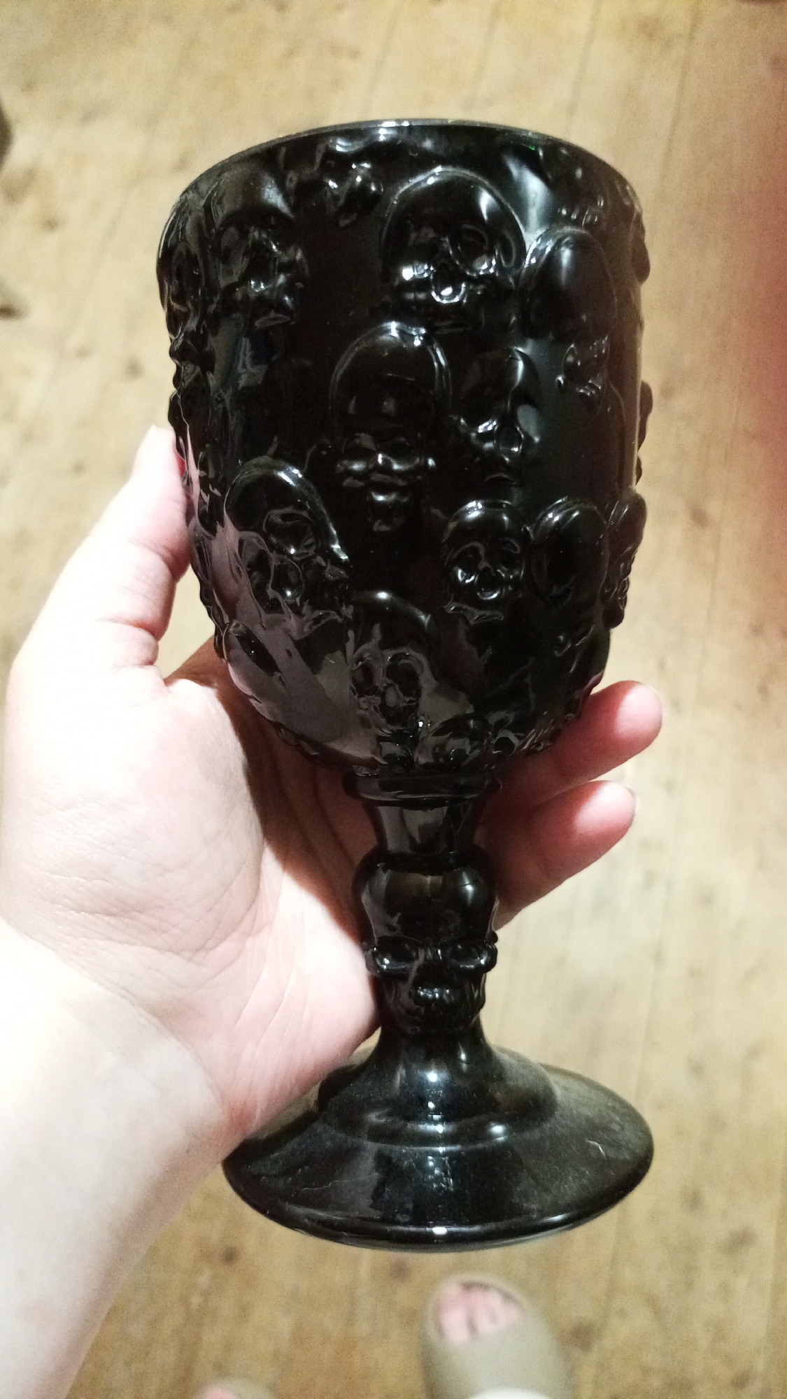 A person is holding a black glass goblet decorated with raised skull designs. The goblet has a wide bowl and a pedestal base. The background shows a light-colored wooden surface. A foot wearing a slipper is partially visible in the bottom of the frame. The location is purported to be Юдино, Russia.