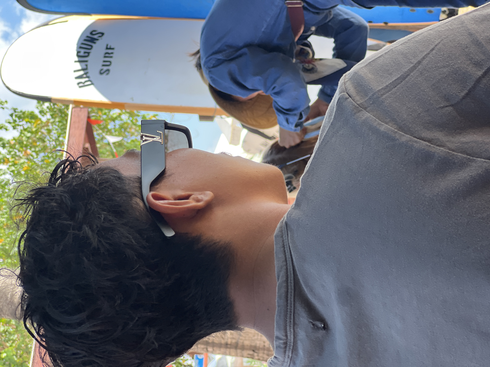 The image depicts a person from the neck up, wearing a grey t-shirt and dark sunglasses with a prominent gold "LV" logo on the side. The person has dark, curly hair. In the background, two surfboards are visible, one white with "BALIGUNS SURF" written on it in green lettering, and another blue surfboard. To the right of the frame, another person wearing a blue shirt and a hat is visible, appearing to be attending to something or someone. The environment suggests an outdoor setting, possibly near a beach, with foliage and a bright sky visible. The overall impression is a casual, possibly vacation-related scene in Kuta, Indonesia.