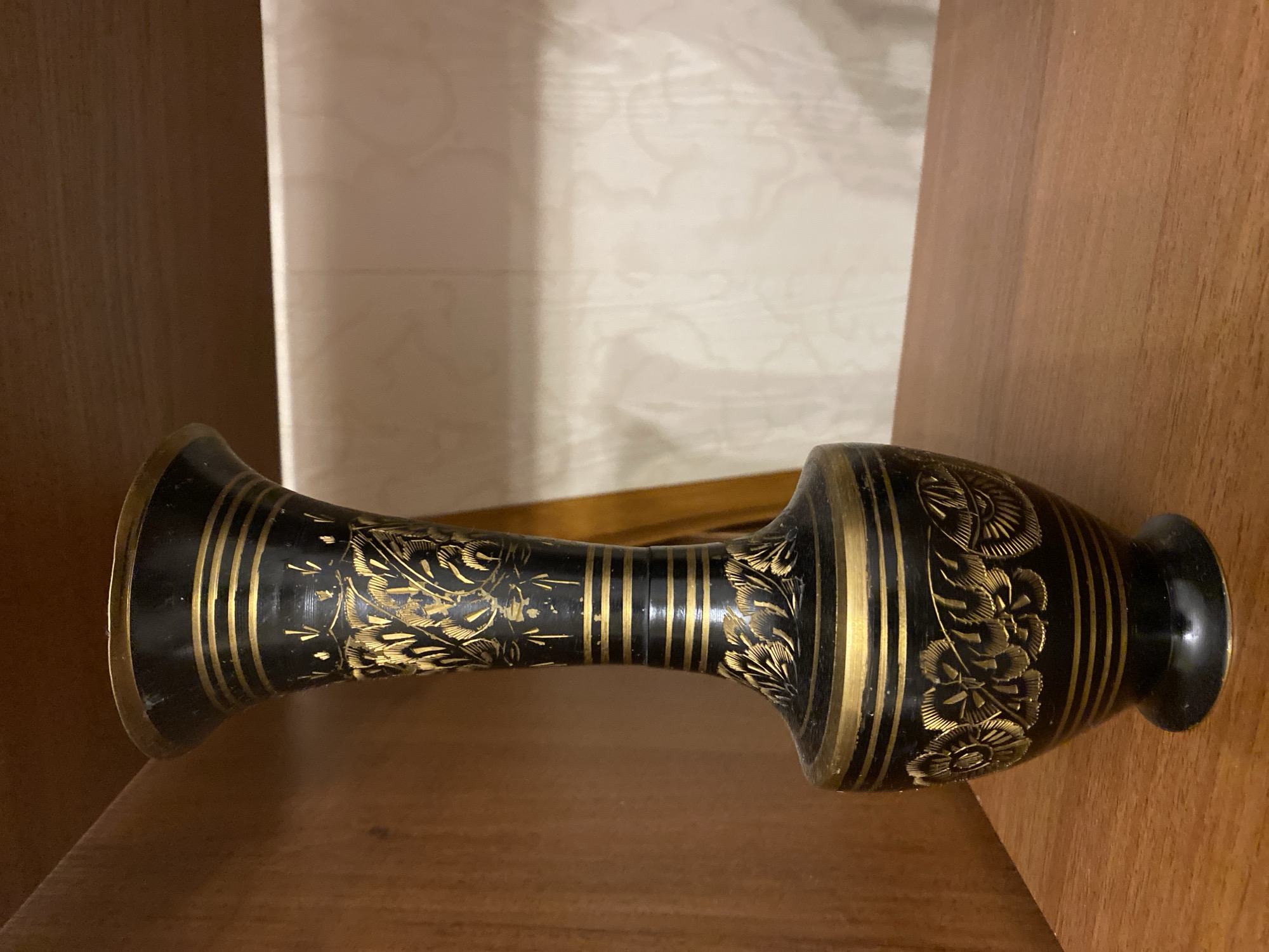 An ornate black vase with extensive gold detailing is positioned on a medium-brown wooden shelf. The vase features a flared top rim, multiple horizontal gold bands, and intricate gold floral and foliage patterns across its upper and lower body sections. The central neck section is narrower and also adorned with gold bands and small vertical dashes. The vase rests on a small, circular black base. Above and to the left, another wooden shelf is visible. To the right of the vase, a light-colored wall with a subtle texture or pattern is present, illuminated by an unseen light source. No individuals are present. The scene is located in Unknown, Unknown.