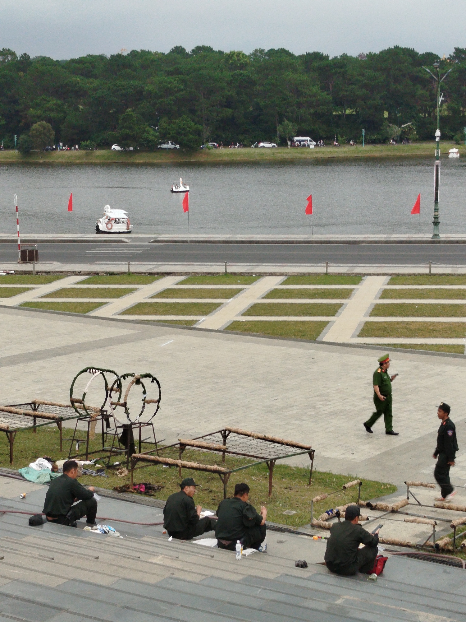 The image shows a wide view of Xuan Huong Lake in Dalat, Vietnam. The lake is calm and grey, reflecting the overcast sky. On the far side of the lake, a dense forest of green pine trees lines the shore. In the foreground, a paved plaza with geometric grass inlays is visible. Several individuals are present in the scene.
In the middle ground, near the lake's edge, a white swan-shaped paddle boat with a red life preserver is docked. Further out on the lake, another white paddle boat is visible. Two red flags are planted on poles in the water near the shore, one on each side of the swan boat.
On the paved plaza, two men in dark green uniforms are walking. One man, wearing a cap, is walking towards the right of the frame, while the other, with a military-style cap and darker uniform, is walking towards the left. Several other men in dark green uniforms are seated on tiered steps in the foreground, facing away from the camera. Some appear to be eating or resting. In the background, on the grass area between the paved plaza and the lake, there are several low-lying metal structures with netting and rope, possibly for decoration or a temporary setup. Litter is visible on the grass near these structures. A street runs along the edge of the lake, with a few vehicles parked on the far side.