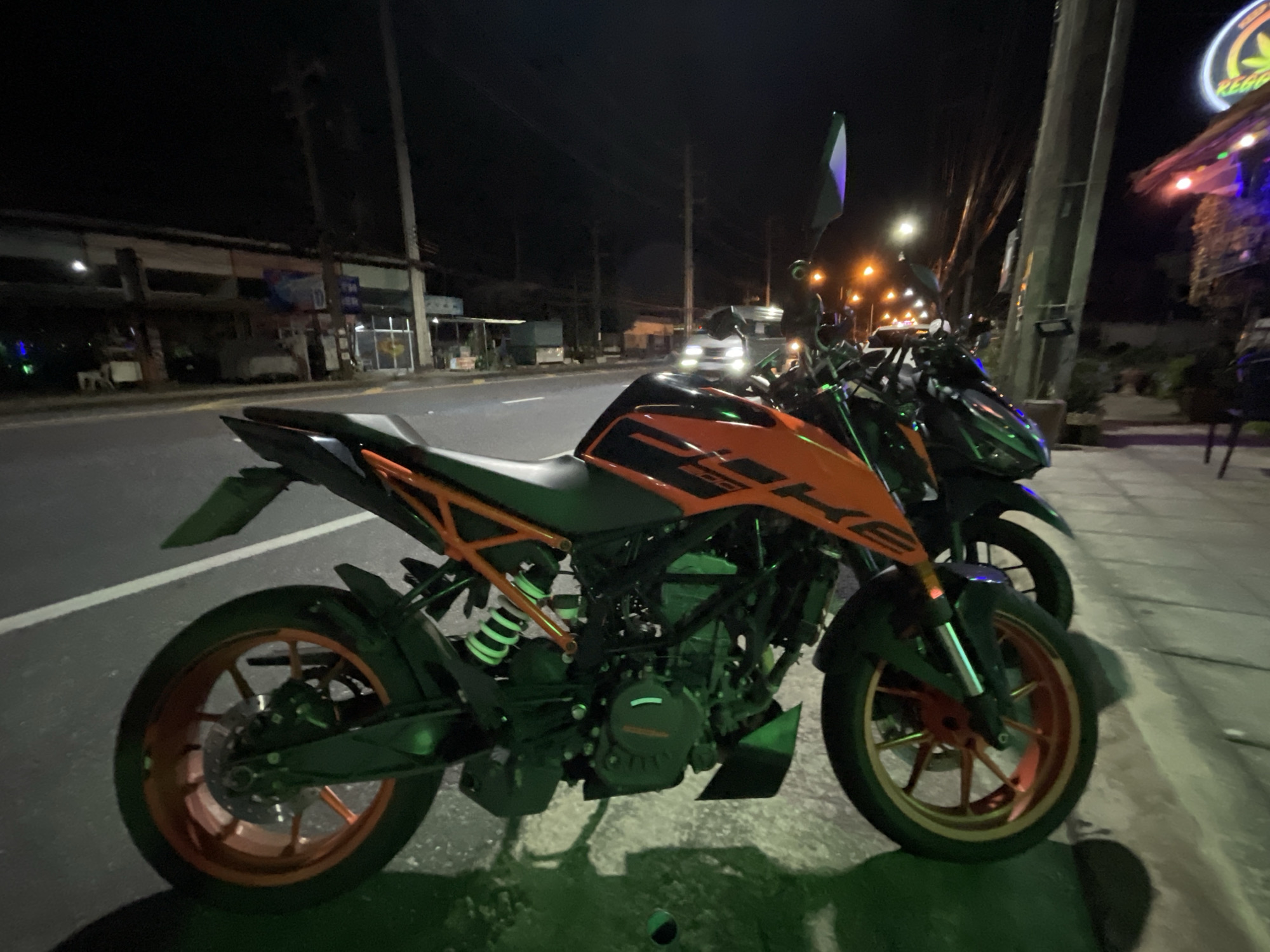 This image depicts two motorcycles parked on the side of a street at night in Chalong, Thailand. The motorcycle in the foreground is an orange and black KTM sportbike with bright orange rims on both wheels. The rear wheel and suspension are clearly visible, as is the engine block. The front of the motorcycle is also visible, showing its wheel and front suspension. Behind it, another motorcycle, primarily black with green accents, is partially visible. The street is paved and has a white line marking the edge of the road. In the background, a building with illuminated signs and colorful lights is visible on the right side of the image. The building on the left appears to be a shop or restaurant, with lights on inside. Streetlights and other ambient lights illuminate the scene, casting shadows on the pavement. The overall environment suggests an urban or semi-urban setting at night, likely a commercial or entertainment area given the illuminated building and the presence of motorcycles. There are no people visible in the image.