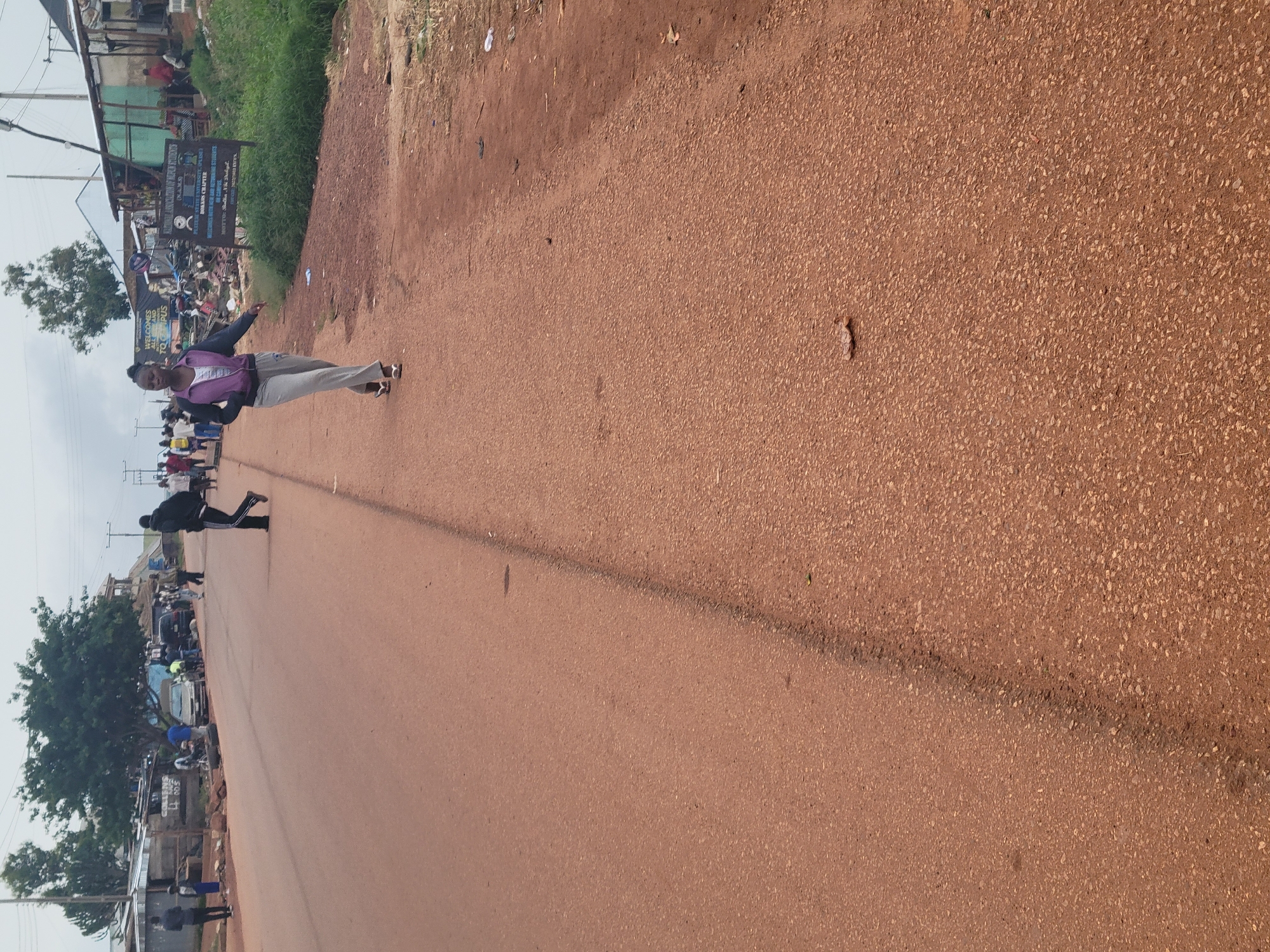 An outdoor, medium shot captures a wide, reddish-brown asphalt road in an urban environment. The road is flanked by a grassy verge and a row of informal structures, including shacks and small buildings, with a few trees visible in the background. The sky is overcast.
In the foreground, a young Black woman with her hair pulled back is walking on the right side of the road, facing left. She is wearing a grey hoodie with a purple jacket over it and grey sweatpants, along with white flip-flops. Her left arm is extended slightly to the side, and her right arm is bent at the elbow.
Further down the road, on the left side, several people are gathered. Some are standing, and others appear to be interacting. A few vehicles, including a silver sedan and a dark SUV, are parked or moving on the road. One man, wearing a dark hooded jacket and black pants with white stripes, is walking away from the camera on the left side of the road.
A sign on the right side of the road displays text that includes "NATIONAL ASSOCIATION OF NIGERIAN STUDENTS" and "PLATEAU STATE UNIVERSITY, OPPS" in English. The overall impression is of a bustling, everyday scene in a developing urban area.