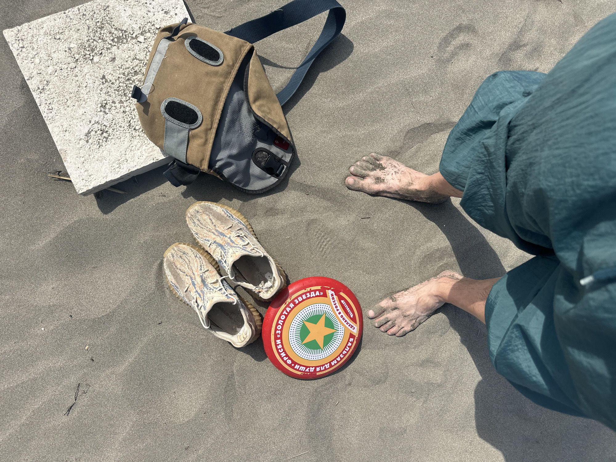 The image depicts a beach scene, possibly in or near Donji Štoj, Montenegro, given the presence of sand. A person is seated, visible from the knee down, wearing teal-colored pants. Their feet are sandy. Nearby are a pair of light-colored sneakers, a red and white frisbee with Cyrillic text and a green star, and a tan and gray messenger bag. A white, concrete-like block lies to the side. The environment suggests a casual beach setting.