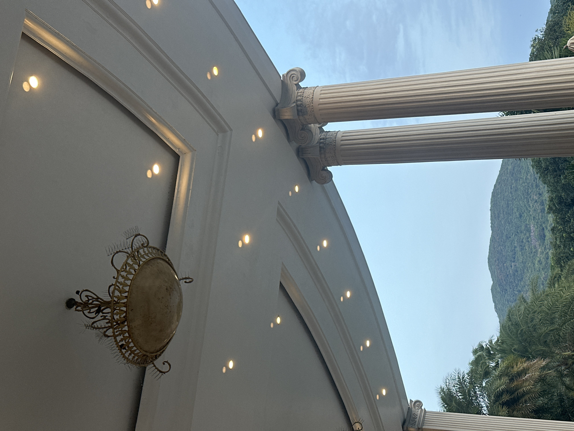 The image captures a low-angle view of a building's exterior, potentially a grand hotel or resort, situated in an area with lush vegetation. The primary architectural elements include classical-style columns with ornate capitals, suggesting Greek or Roman influences. A curved, white-painted ceiling is visible, embedded with evenly spaced recessed lights that create symmetrical patterns. A decorative, circular light fixture with an intricate metal frame adorns the ceiling. Outside the building, a background of dense greenery and a mountainous landscape is visible, set against a pale blue sky. The scene is tranquil, likely depicting a daytime setting in a tropical or subtropical environment. The location is Unknown, Unknown.