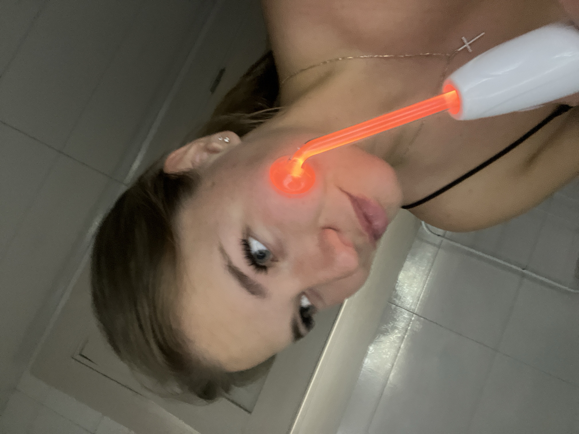 A young woman is captured in an indoor, dimly lit setting, seen from a high angle. The image is rotated approximately 90 degrees counter-clockwise. She has light skin, striking blue eyes, and pronounced eyelashes and eyebrows. Her mouth is slightly ajar, suggesting focus or a relaxed expression.
She is engaged in a skincare activity, using a high-frequency facial device. The device has a white handle, and its glass electrode tip, shaped like a mushroom, emits a bright, glowing orange light as it touches her left cheek. This glowing element is the primary light source in the frame, casting a warm hue on her face.
Around her neck, she wears a delicate gold chain with a small white cross pendant. A dark strap, likely from a top or undergarment, is visible over her right shoulder. Her hair, appearing dark brown, is styled away from her face on the visible side.
The background consists of light-colored, possibly white or grey, tiled walls with visible grout lines, hinting at a bathroom or a clinical/spa environment. The overall ambiance is dark, with the device's glow providing localized illumination. No text is visible in the image. This scene takes place indoors in Budva, Montenegro.