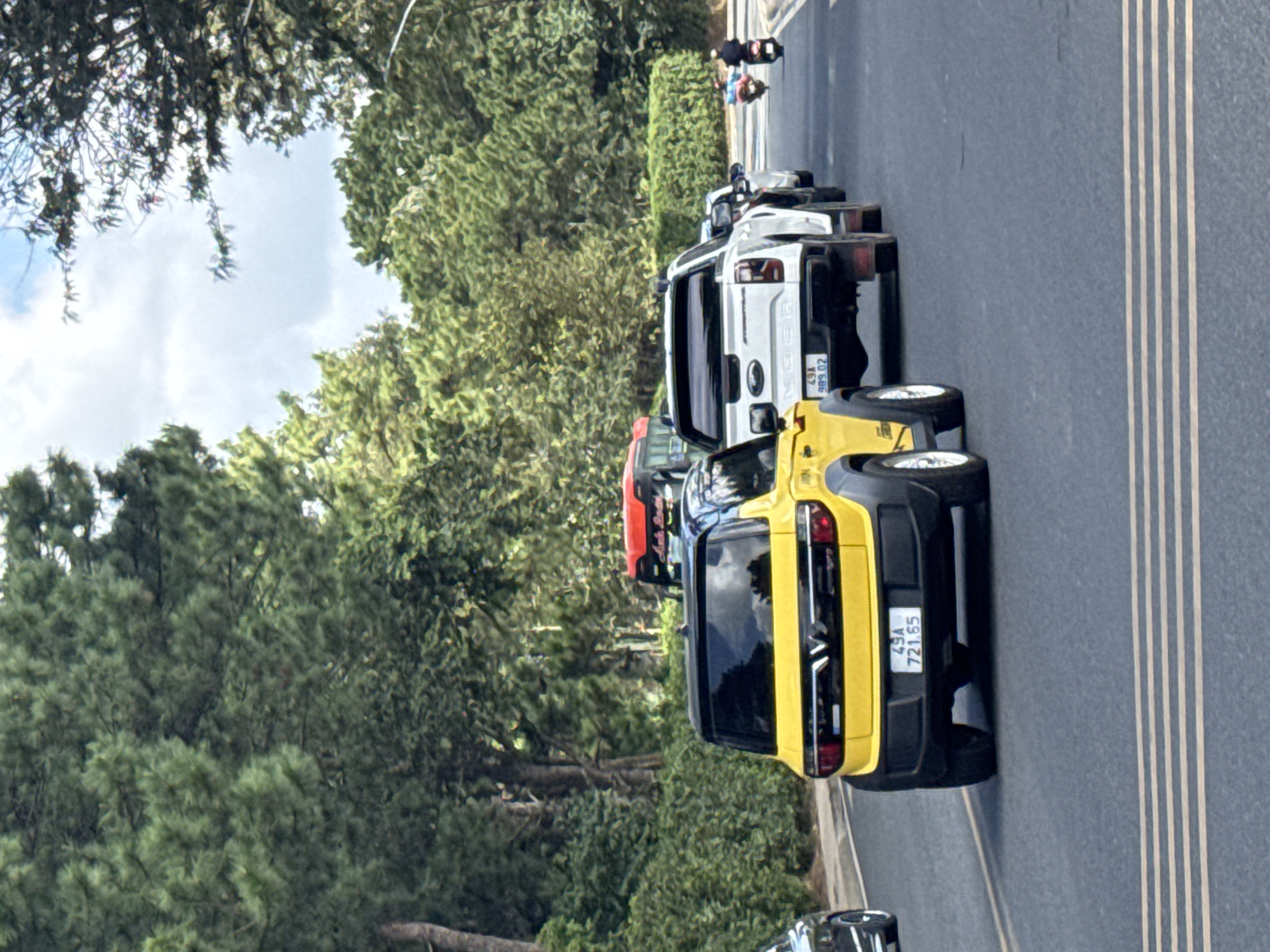 In Da Lat, Vietnam, a yellow car with license plate "49A 721.65" is parked on a road lined with lush green trees and bushes. A white pickup truck with license plate "49A 989.02" follows closely behind the yellow car, also on the road. Further down the road, two individuals are seen riding motorcycles. The sky above is partly cloudy. The scene appears to be a typical day on a road in Da Lat, characterized by its natural beauty and busy traffic.