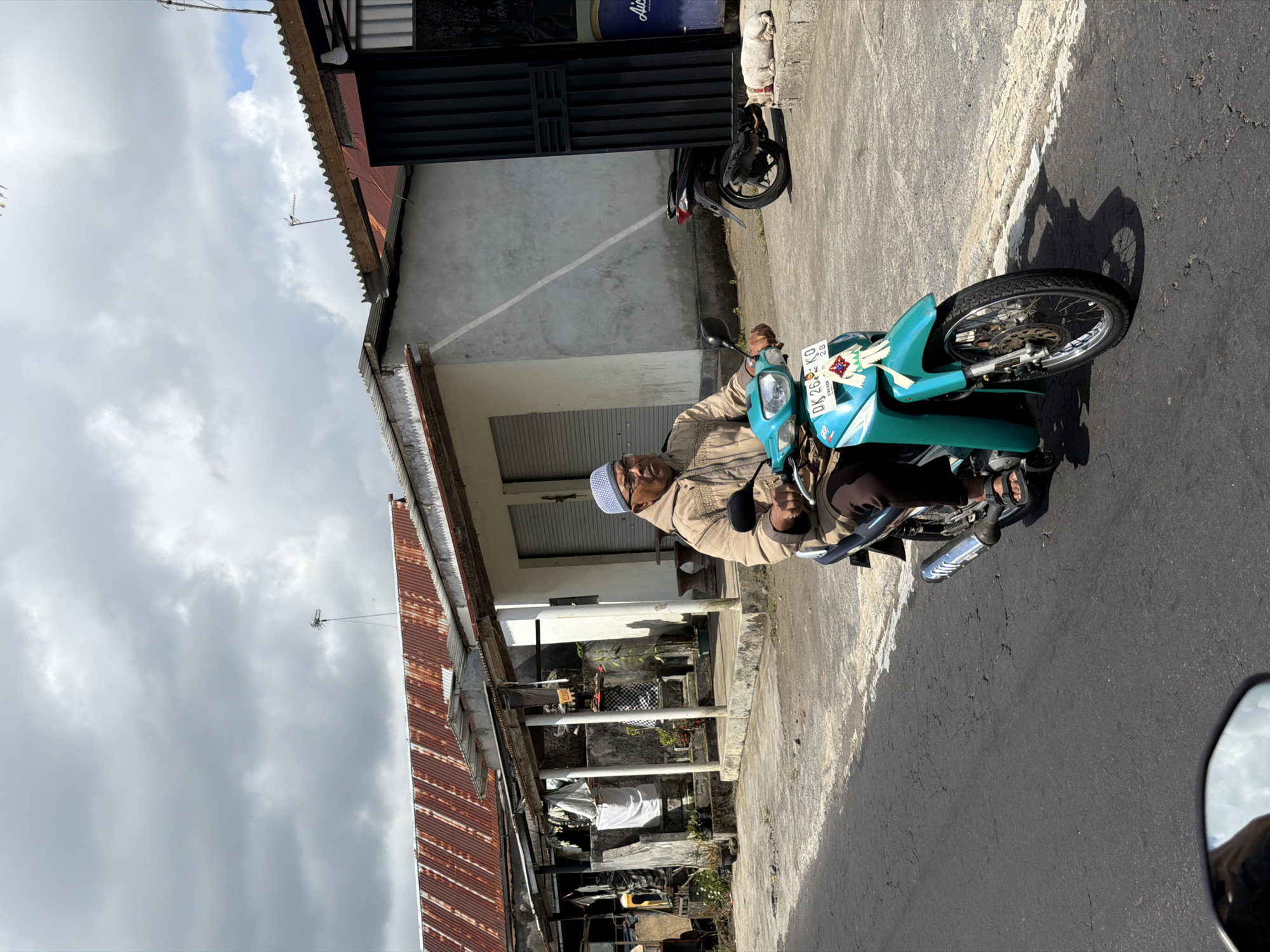 A man wearing a light brown jacket and a white skullcap rides a turquoise motorcycle down a cracked asphalt road in Kintamani, Indonesia. He is wearing glasses and dark pants, and his feet are in sandals. The motorcycle has a license plate that reads "DK 268 KO". In the background, there are buildings with corrugated metal roofs and a white wall with a dark metal gate. A white dog is lying on the ground near the gate. The sky is cloudy.