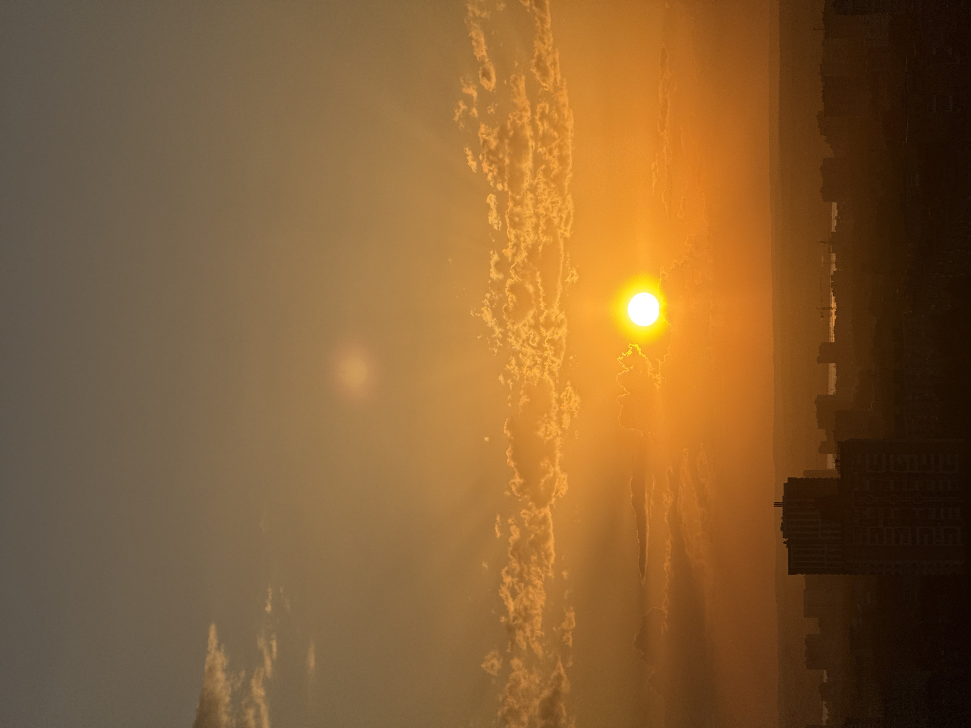 The image captures a sunset over a cityscape, likely Shirkovo, Russia. The sun is a bright orb, partially obscured by clouds that stretch vertically across the center of the frame. Beams of light radiate from the sun through gaps in the clouds. To the right, the silhouettes of buildings are visible against the bright sky, suggesting an urban skyline. The overall color scheme is dominated by orange and yellow hues, typical of a sunset. The sky transitions from a muted gray on the left to a vibrant orange near the sun. A second, fainter circular area of brightness is present in the sky to the left of the sun.