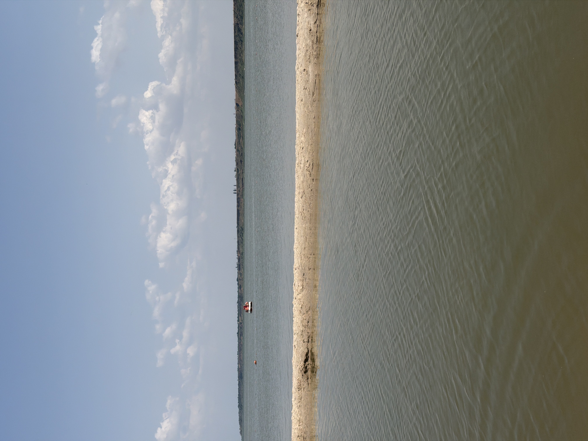 The image depicts a serene water scene, likely a bay or river, in Pavlo-Ochakovo, Russia. In the foreground, a narrow strip of sandy land juts out into the water, creating a visual division. The water itself is a muted green-brown, with subtle ripples indicating a gentle current. In the middle ground, on the surface of the water, a small red and white vessel is visible, possibly a boat or a floating structure. Further in the distance, a faint line of land stretches across the horizon, with some vegetation and possibly buildings dotting the landscape. The sky above is a pale blue, with scattered white, puffy clouds. The overall atmosphere is peaceful and natural, suggesting a calm day in a coastal or riverside environment.