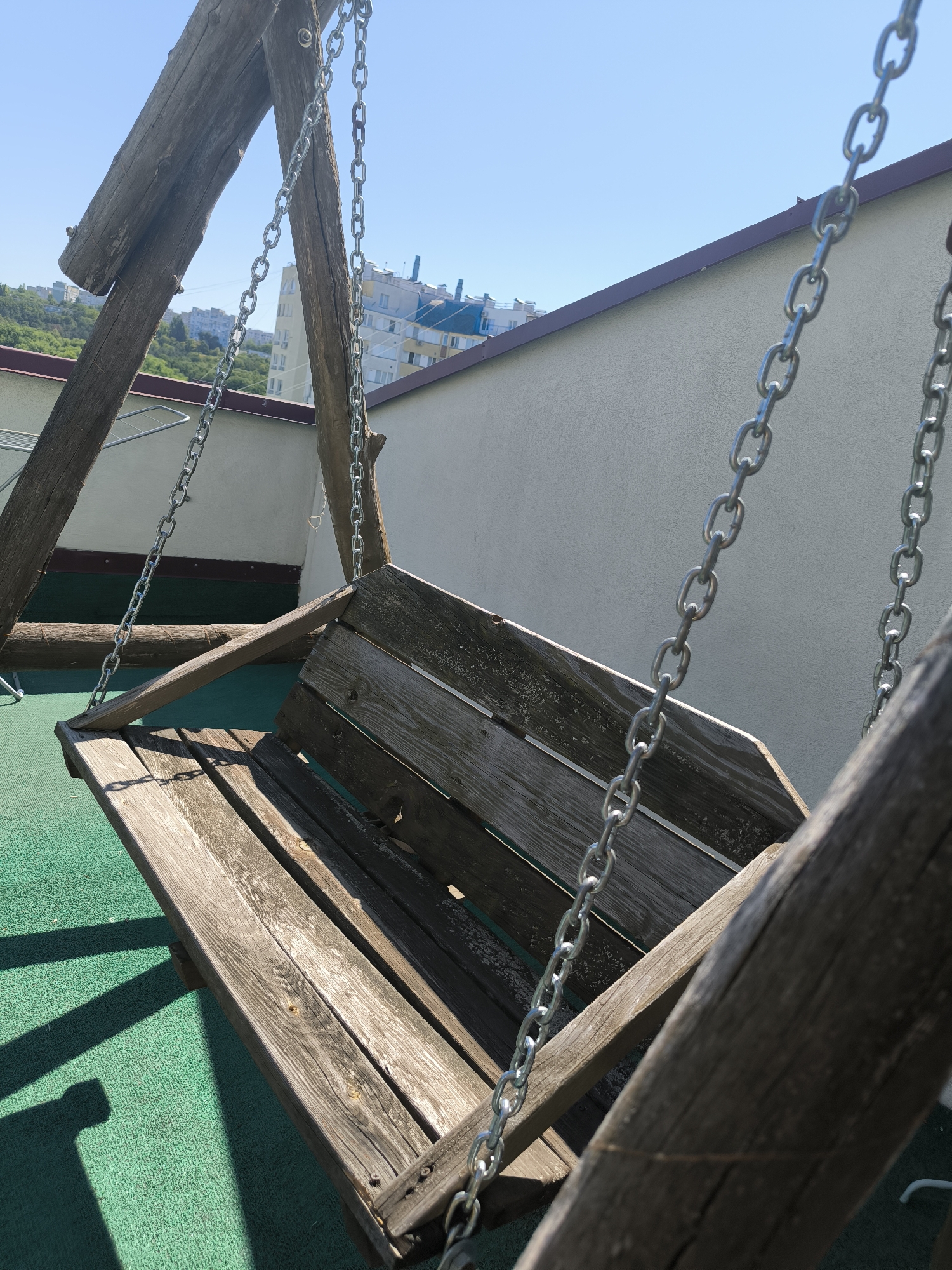 An outdoor wooden swing set with a weathered wooden bench is situated on a green, textured surface. The swing is suspended by metal chains. In the background, a light-colored building with a dark-brown roofline is visible, and beyond that, several multi-story buildings typical of an urban environment can be seen. The sky is clear and blue. This scene appears to be a rooftop or elevated balcony area in the city of Chișinău, Moldova, based on the architectural style of the background buildings. There are no people or animals present in the image. The lighting suggests it is daytime, likely morning or afternoon, given the presence of shadows.
