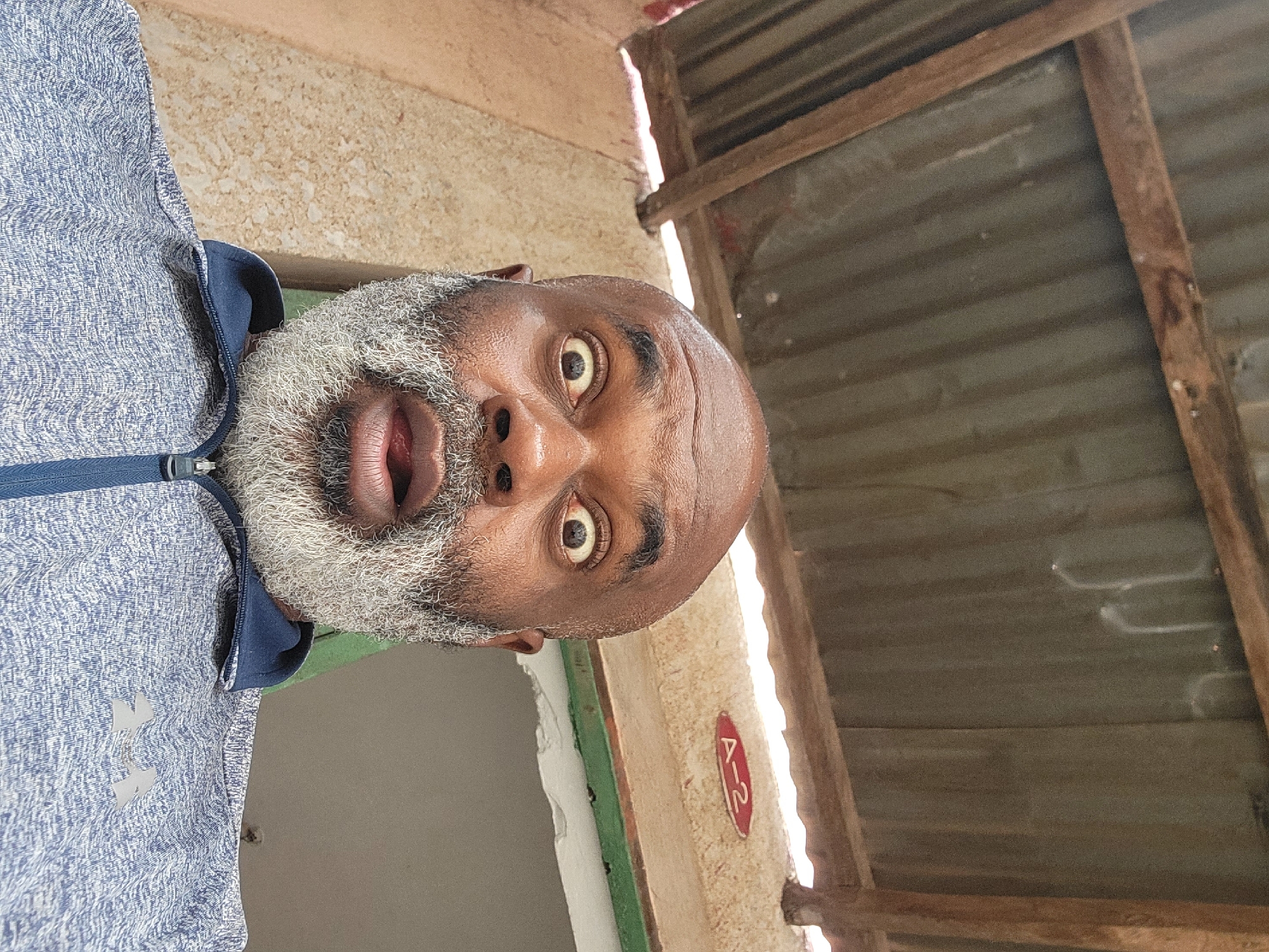 A selfie of a Black male with a bald head and grey beard, wearing a blue and white patterned zip-up jacket with a navy collar, taken from a low angle. The man is looking directly at the camera with a surprised expression. The background shows a building interior/exterior, featuring a corrugated metal roof with wooden supports and a light beige wall. A partially visible doorway, painted green and white, is on the left. A sign with "A-2" in red is affixed to the wall. The location is likely Maiburiji, Nigeria, based on the prompt.