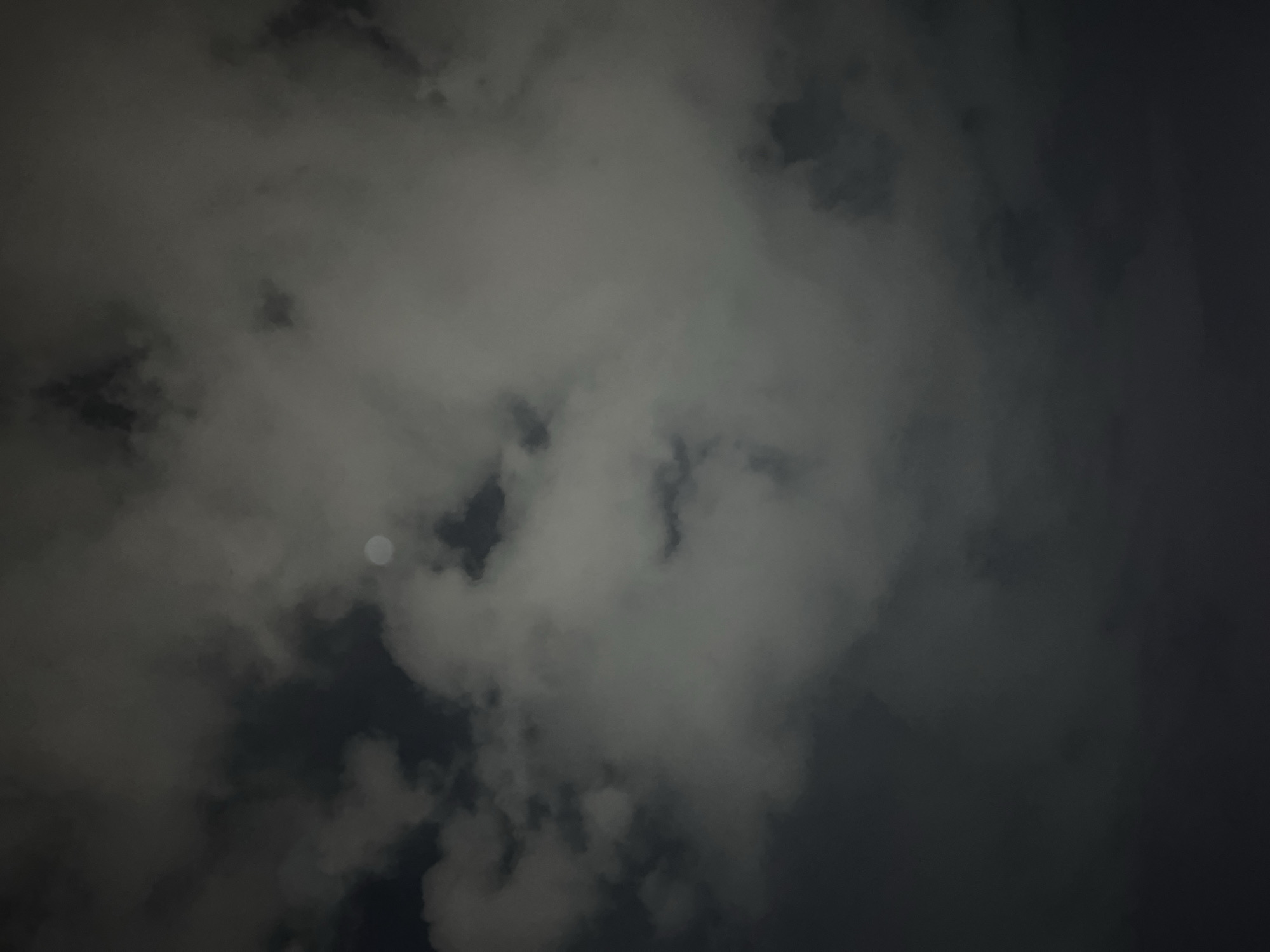 The image depicts a night sky filled with dark, heavy clouds, obscuring most of the view. A small, pale moon can be seen peeking through a gap in the clouds. The scene is dark and overcast. There are no identifiable people, subjects, or actions visible. The location, Yerevan, Armenia, cannot be verified from the image.