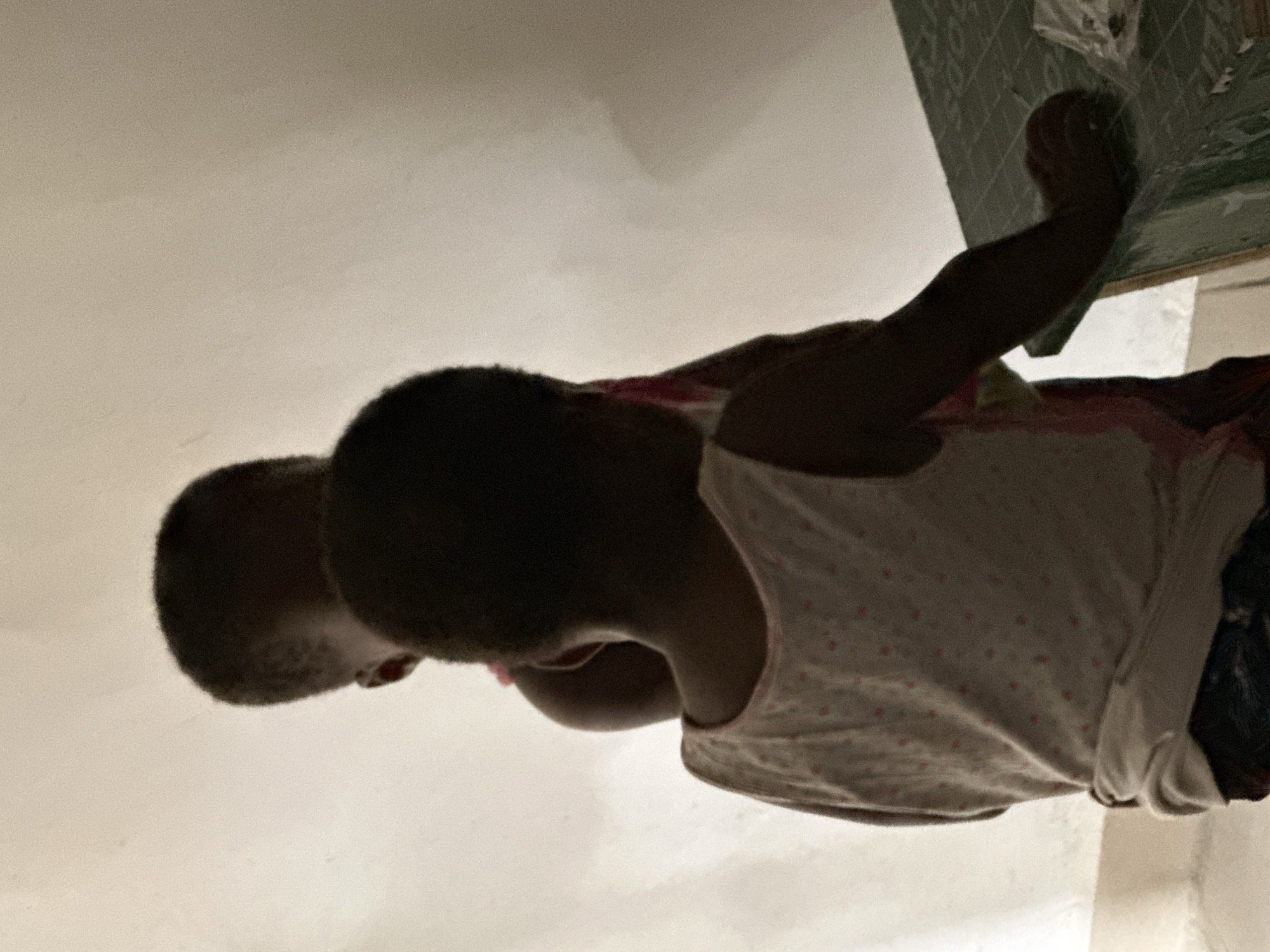 In a room in Narayi, Nigeria, two children are seen from behind, silhouetted against a brightly lit wall. The child in the foreground, wearing a light-colored tank top with small pink dots, is positioned to the left of the frame, with their back to the viewer. This child's head is tilted downwards, and their left arm is bent, resting on a green surface to the right. A portion of a patterned cloth or fabric is visible near their shoulder. Behind and slightly to the right of the first child is another child, also seen from behind, whose head is positioned higher. This second child appears to be wearing a pink garment underneath their outer clothing. The environment is simple, with a plain, illuminated wall dominating the background and a glimpse of a patterned mat or table surface in the foreground. The lighting suggests the image was taken indoors, possibly during daytime, with a strong light source behind the children.