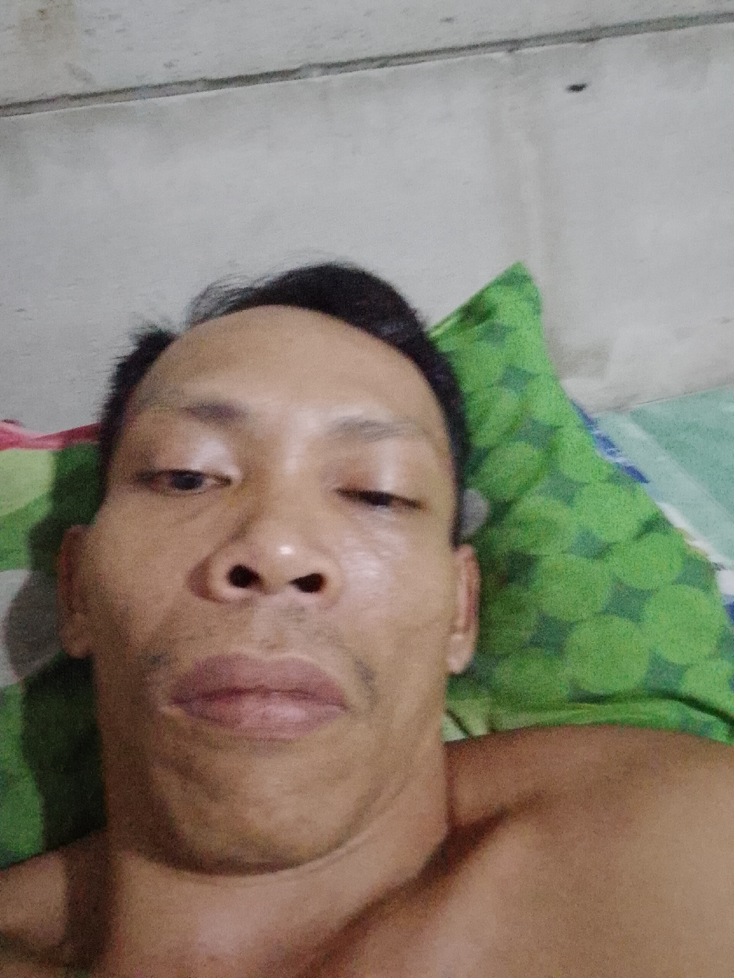 A selfie-style image of a man lying on a patterned bed. He appears to be indoors with a wall visible in the background. The location is Bukit Ketok, Indonesia.