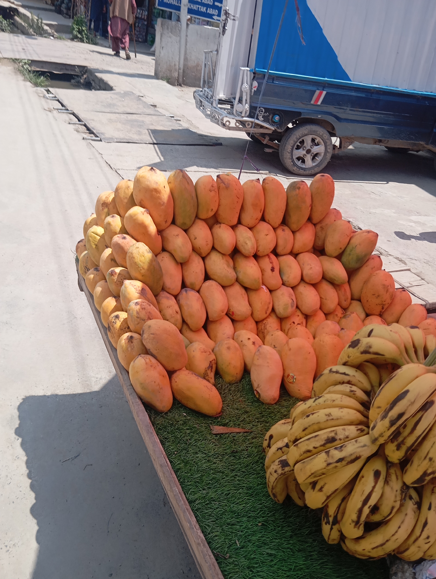 An outdoor scene captures a fruit stand laden with ripe mangoes and bananas. The mangoes, predominantly orange and yellow, are artfully stacked on a wooden display covered with artificial green turf. A bunch of yellow bananas with brown spots is placed to the right of the mangoes. In the background, a blue and white truck is parked, and a person in a traditional outfit walks away from the camera. The ground is paved with concrete, and the sunlight casts shadows across the scene, suggesting it is daytime. The location appears to be a street or market.