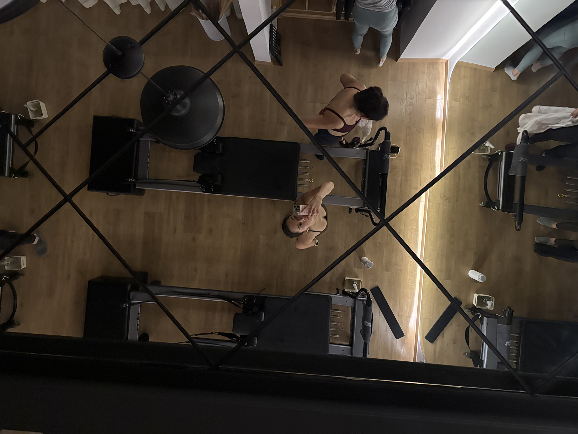 A high-angle shot, possibly taken through a lattice-like structure, reveals an exercise studio in Puebla, Mexico. The studio has light-colored wooden flooring. Visible are several Pilates reformers, complex exercise machines with sliding carriages, springs, and straps. Two women are present, one is using a reformer while the second one is photographing herself in the mirror while standing next to a reformer. A third person is also visible, partially obscured in the upper right corner of the frame, apparently observing the exercise session. The ceiling has recessed lighting that casts light into the studio. Water bottles are scattered on the floor.