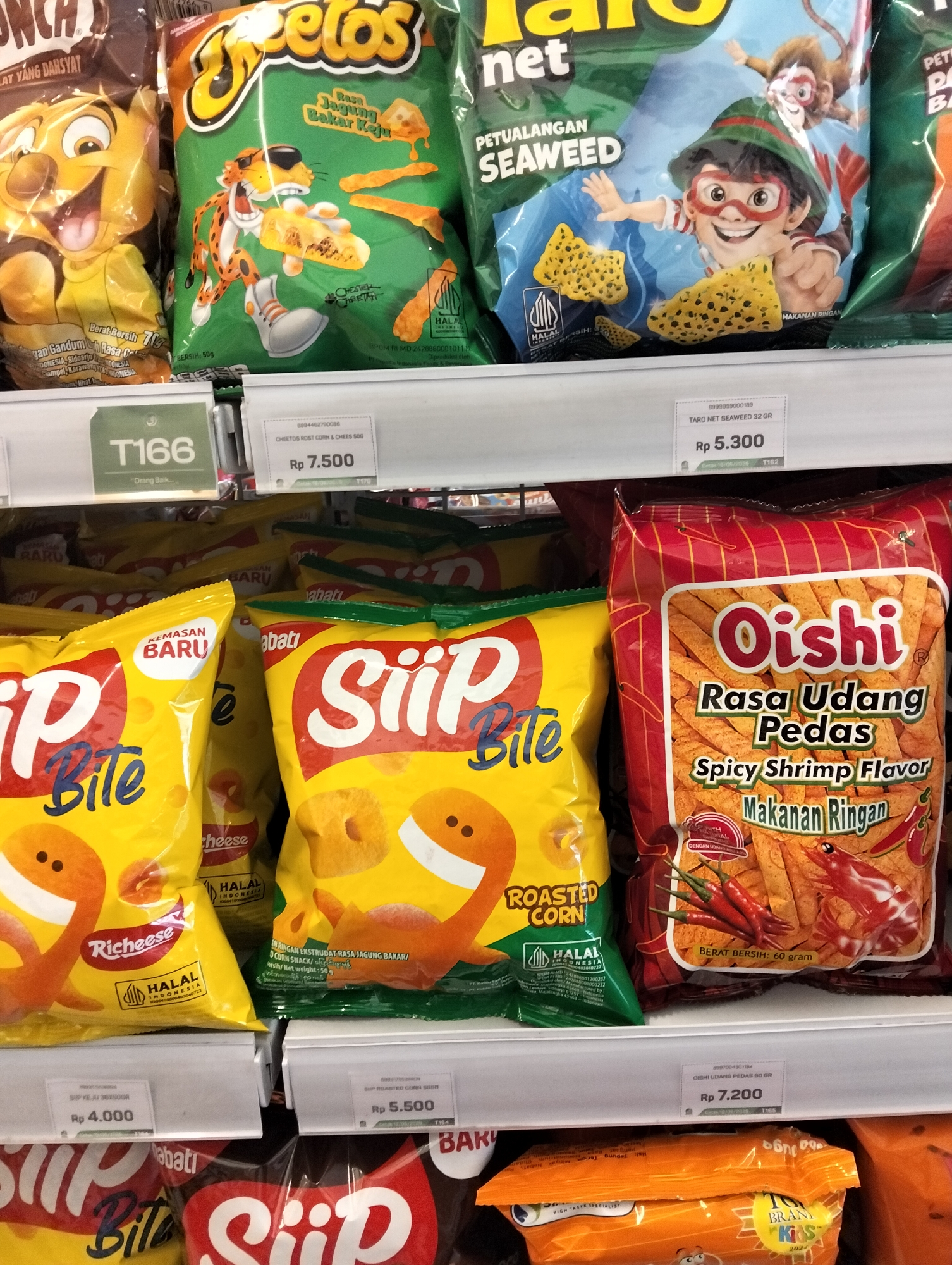 The image shows a retail shelf stocked with various snack food packages. Dominant brands include Cheetos, Taro Net, Siip Bite, and Oishi. The snacks are displayed in colorful bags, with some featuring cartoon characters and appealing food imagery. Price tags are visible for some items, indicating prices in Indonesian Rupiah (Rp). For example, "Cheetos Rost Corn & Cheese 50G" is priced at Rp 7.500, and "Taro Net Seaweed 32 Gr" is priced at Rp 5.300. "Siip Keju 36X50GR" is priced at Rp 4.000, and "Siip Roasted Corn Dorr" is priced at Rp 5.500. "Oishi Udang Pedas 60 Gr" is priced at Rp 7.200. The arrangement suggests a typical supermarket or convenience store setting. The lighting is bright, illuminating the products clearly. The background shows more rows of snacks on the shelves. The overall scene is clean and organized. The information provided is purely descriptive and does not allow for any conclusions about the specific location of Muncar, Indonesia beyond the price indications in Indonesian Rupiah.
