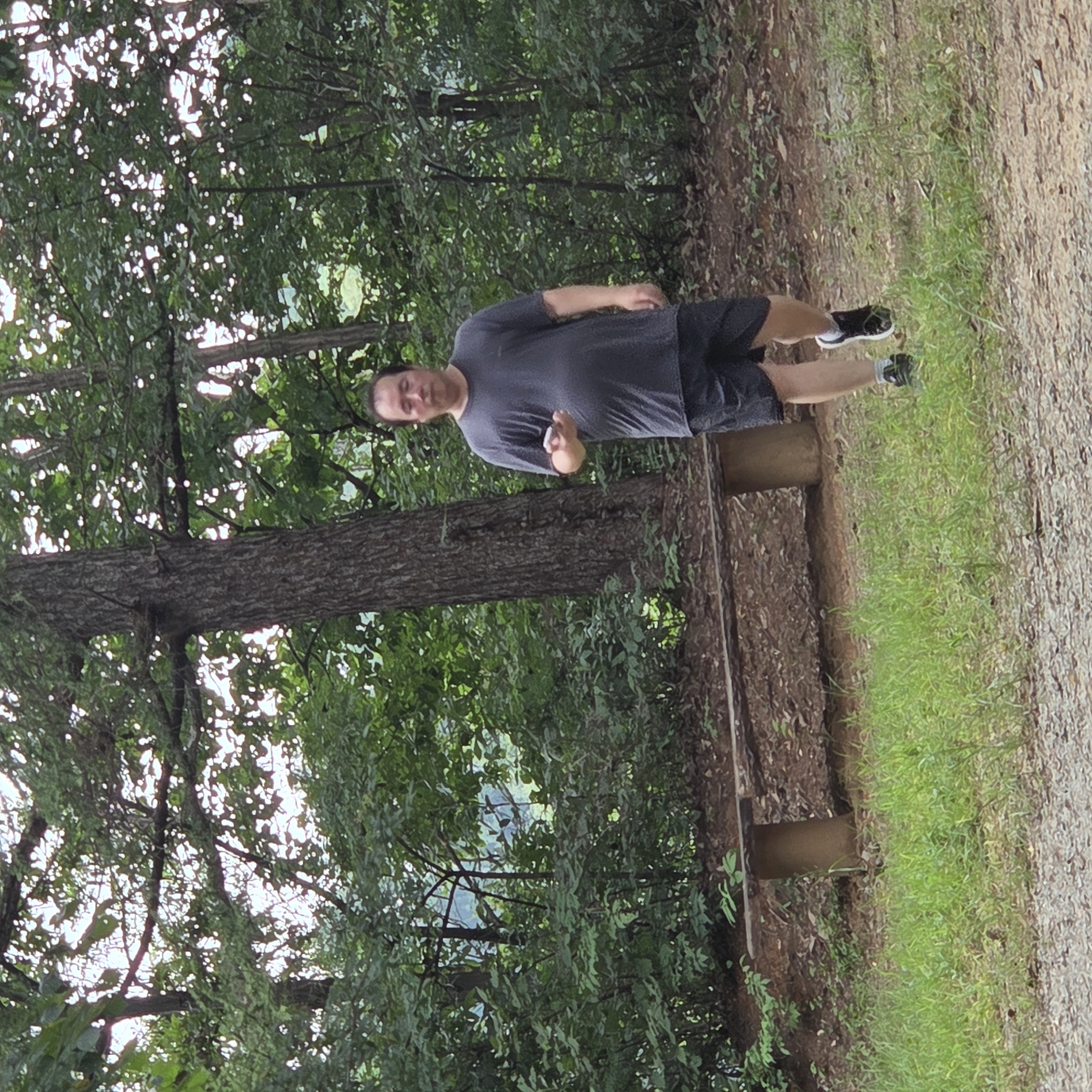 A man is walking towards the camera in a wooded area. He is wearing a dark t-shirt and dark shorts. He is holding a small white object in his right hand. There is a wooden bench to his left, and a large tree behind him. The ground is covered in grass and dirt. The background is filled with lush green trees and foliage. The lighting suggests it is daytime. The location is Gapcheon-myeon, South Korea.