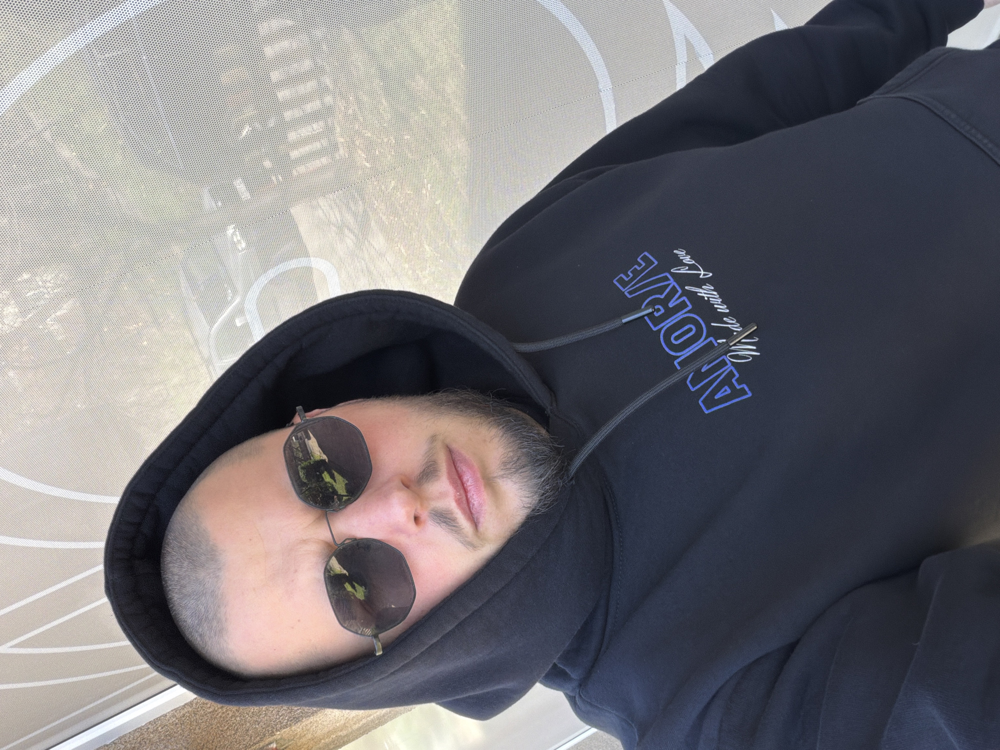 A man wearing a black hoodie and sunglasses is facing the camera. The hoodie has the word "AMORE" in blue lettering with the phrase "We ride with Love" below it. The background consists of a white perforated surface. The location is undetermined.