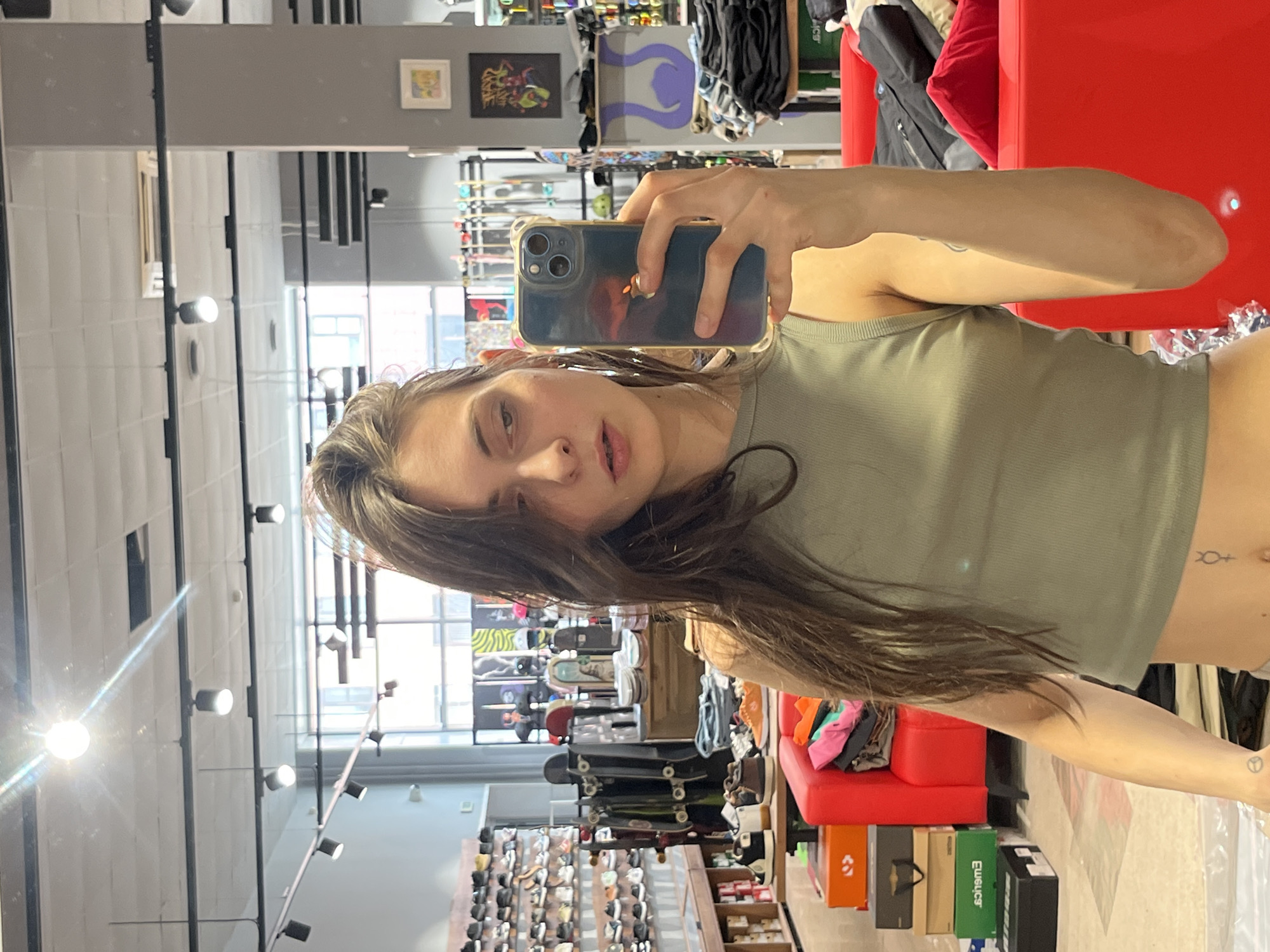 A young woman is taking a selfie in a store that appears to be a skate shop. The store has rows of shoes, skateboards, and apparel. There is a red couch in the background, and boxes are stacked on the floor. The woman is wearing a green tank top and has long brown hair. Her phone is a dark blue model. It is not possible to determine the exact location within the city of Krasnoyarsk, Russia from this image.