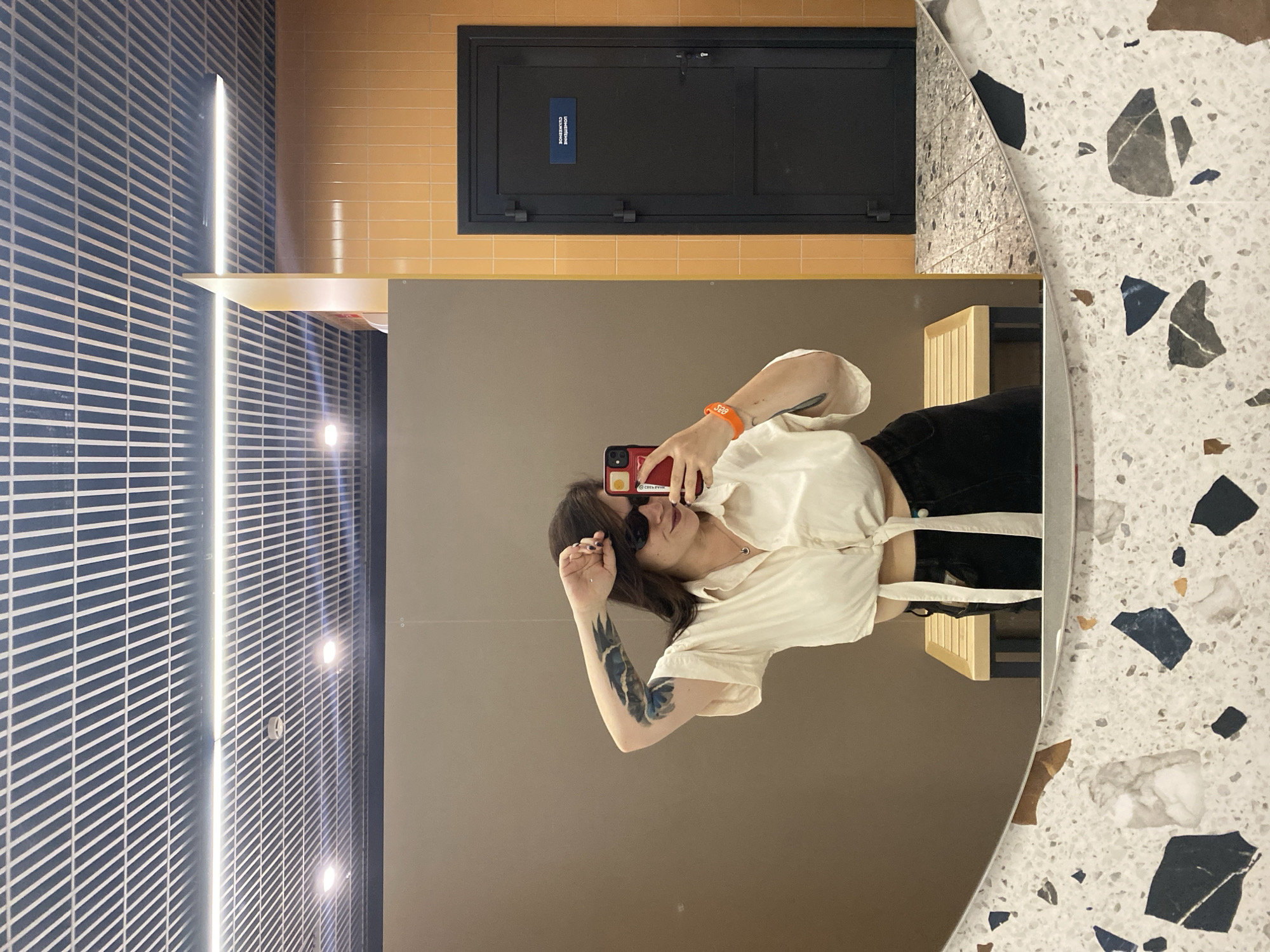A young woman with dark hair and sunglasses stands in a changing room, taking a mirror selfie. She is wearing a cream-colored tie-front blouse and black pants. A tattoo is visible on her left arm. The changing room has a neutral-toned wall, orange tiled walls, a black door with a sign, a wooden bench, and a speckled mirror. The ceiling features a grid-like pattern with fluorescent lighting. The scene is likely in a public facility, such as a gym or a department store changing room in Votkinsk, Russia.