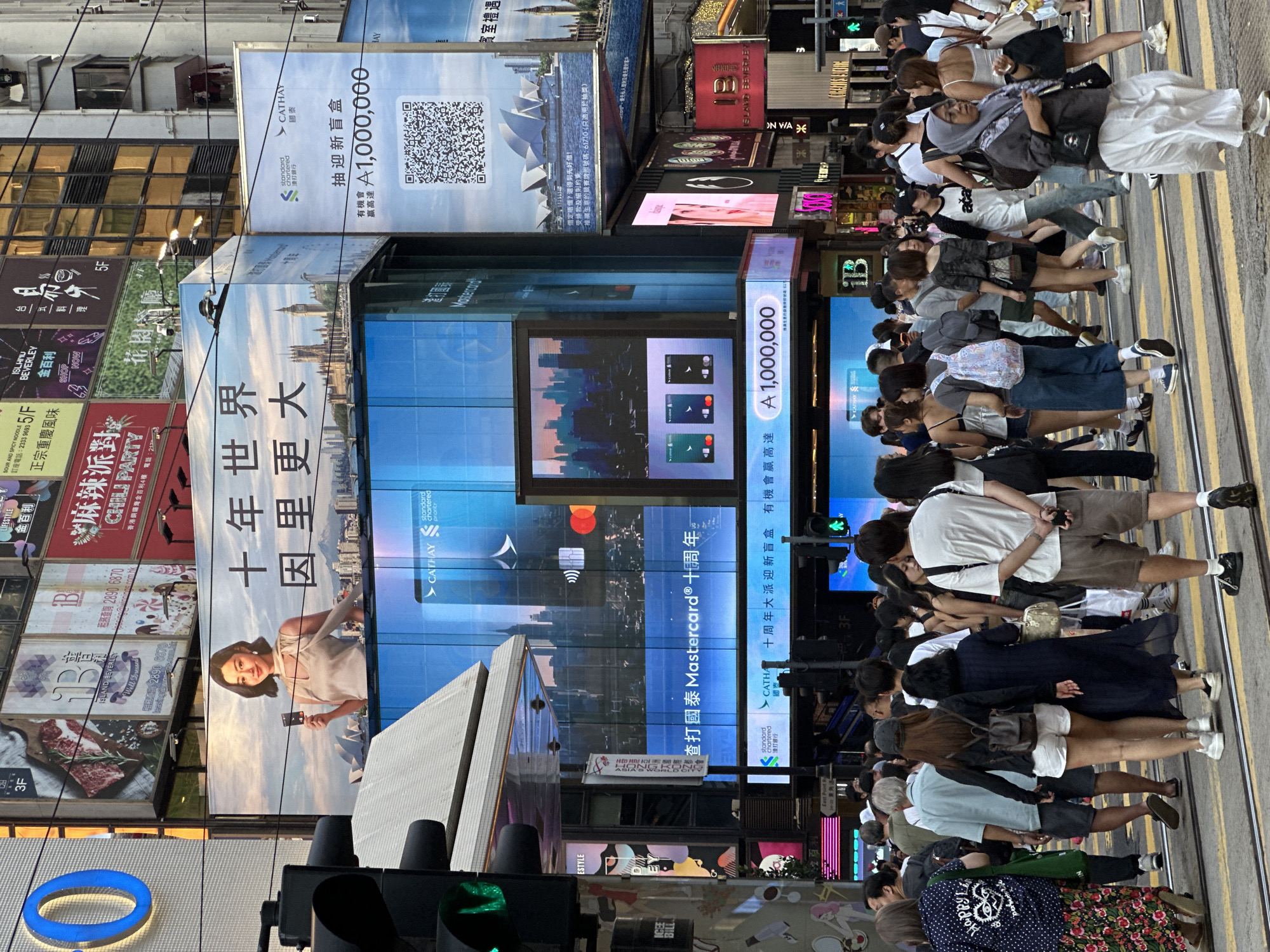 This is a street-level, medium-shot, eye-level, full-color photograph taken on a street in Hong Kong, China. The scene is dominated by numerous large electronic billboards and advertisements mounted on buildings. In the foreground, a crowd of people, appearing to be tourists and locals, are crossing a street marked with yellow and white pedestrian crossings. The people are dressed in casual summer clothing.
Prominently featured are several large digital billboards. One billboard displays a woman holding a credit card, with text in Chinese characters that can be translated to "Ten Years World Bigger". Another large billboard features the logos of Cathay Pacific and Standard Chartered Bank, advertising a prize of "A 1,000,000" and a QR code. Below this, a digital screen shows various credit card designs. Further advertisements show the Sydney Opera House and text in Chinese. Smaller signs and advertisements for businesses like "Island Beverley" and "Chili Party" are also visible.
The environment is urban, with tall buildings, power lines, and traffic signals indicating a busy city intersection. The lighting suggests it is daytime, possibly late afternoon, with a mix of sunlight and the glow from the electronic displays. The overall impression is one of a vibrant and commercial urban center.