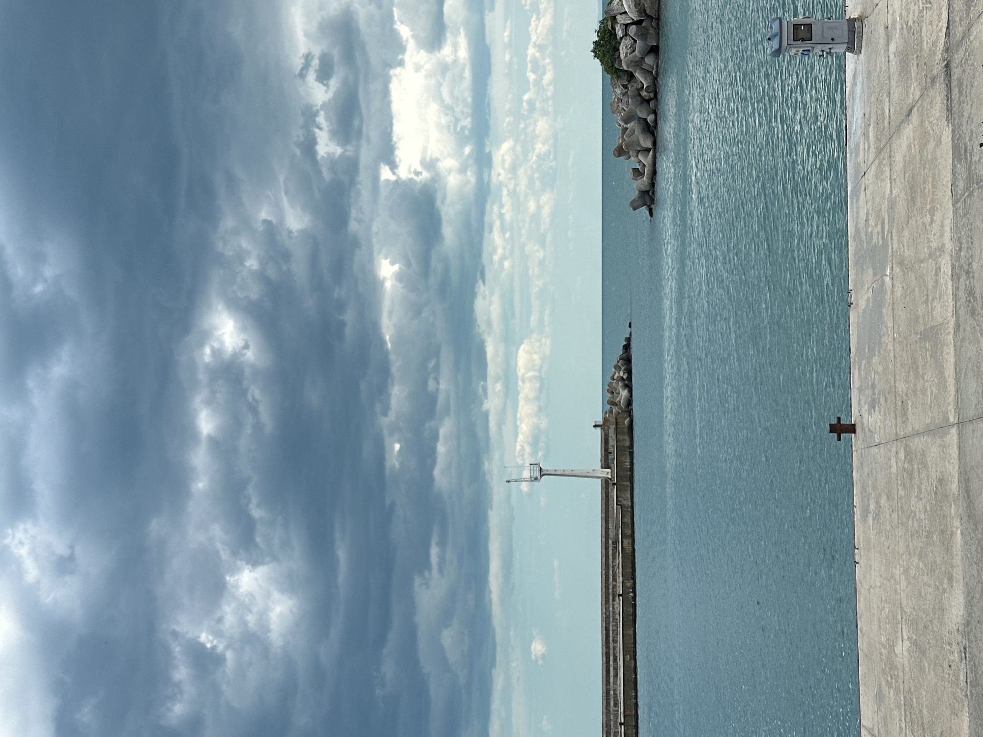 The image shows a view in Varna, Bulgaria, likely along the coast. Dominating the left side of the frame are dense, dark, and dramatic storm clouds. The right side consists of blue-green sea water, a concrete pier, and a smaller rock breakwater. A white structure, possibly a navigational aid or small tower, is positioned along the pier. The pier is grey and appears to be made of concrete or stone. Further out in the sea, there's a collection of large, geometrically-shaped concrete blocks, likely tetrapods, acting as a larger breakwater, with some greenery atop it.