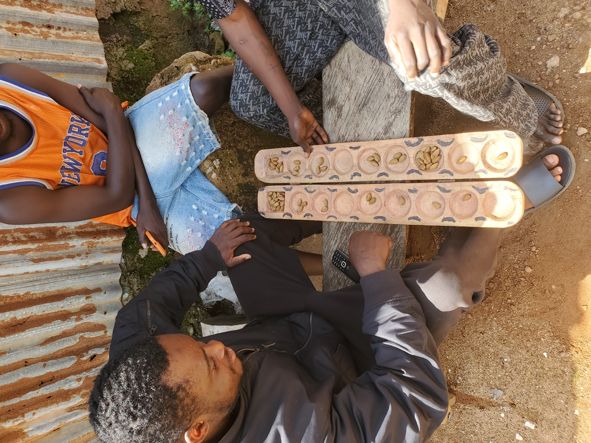 Three individuals are gathered around a traditional Mancala board game, made of carved wood and filled with seeds, possibly during a daytime scene in an outdoor setting in an unknown location in Nigeria. The person on the left, a man with dark skin and a beard, wears a black jacket and is seated, looking intently at the game. He is holding a mobile phone in his right hand. To his right, another individual, partially visible, is also seated and appears to be playing the game. This person is wearing a black and white patterned garment on their lower body. In the background, a third person, a young man, is seated with crossed arms, wearing an orange basketball jersey with "NEW YORK" emblazoned on it and denim shorts. The setting appears rustic, with a corrugated metal sheet as a backdrop and a sandy, dirt ground beneath them. The Mancala board is placed on a wooden surface.