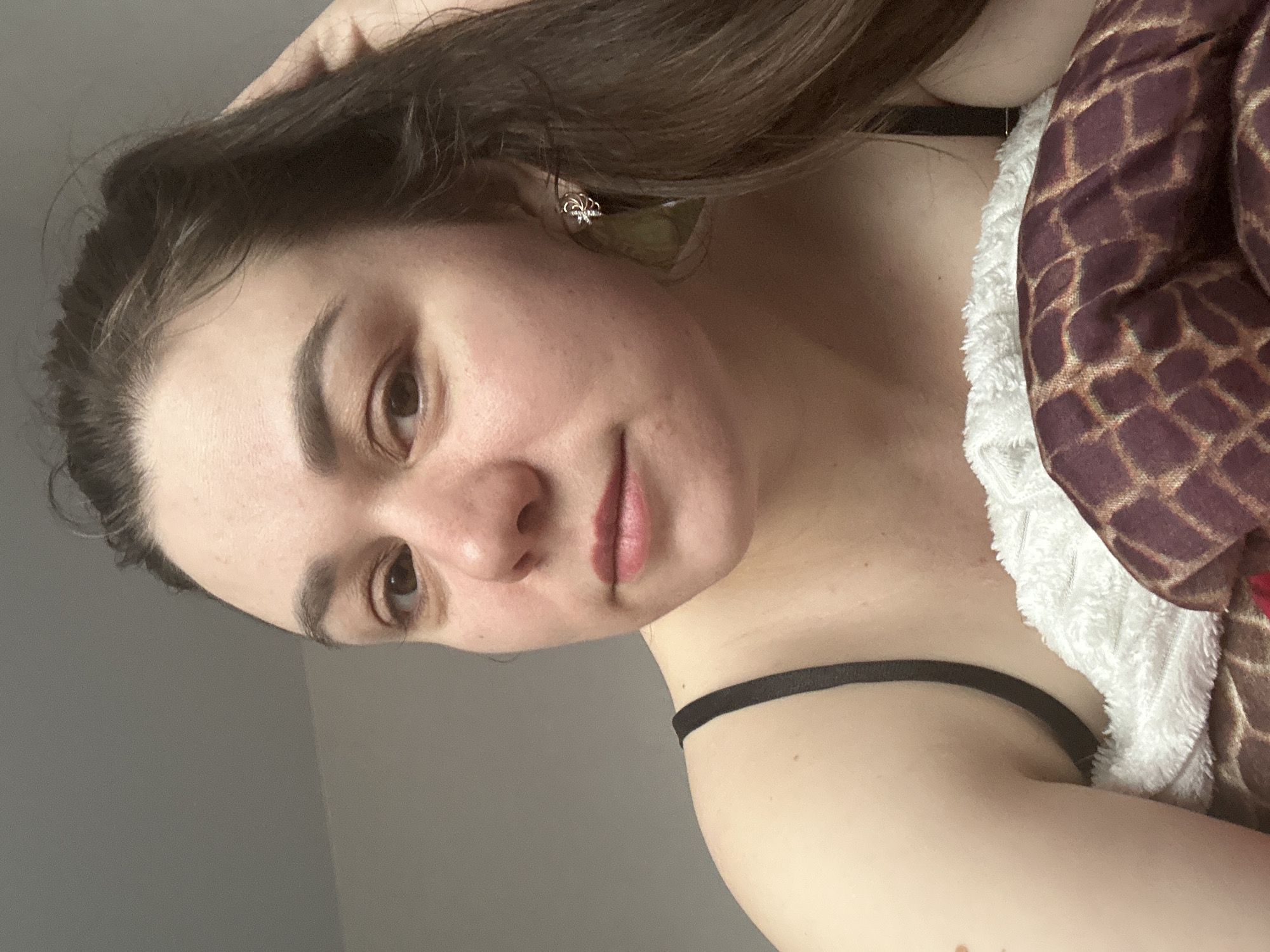 A woman with brown hair and light skin is posing for a photo. She is wearing a black strap, possibly of a top or a dress. She has an earring on her left ear. Her hair is partially covering her face. There is a patterned blanket or garment next to her. The background appears to be a plain wall, likely indoors. There is no indication of the city of Chaikovsky, Russia in the image.