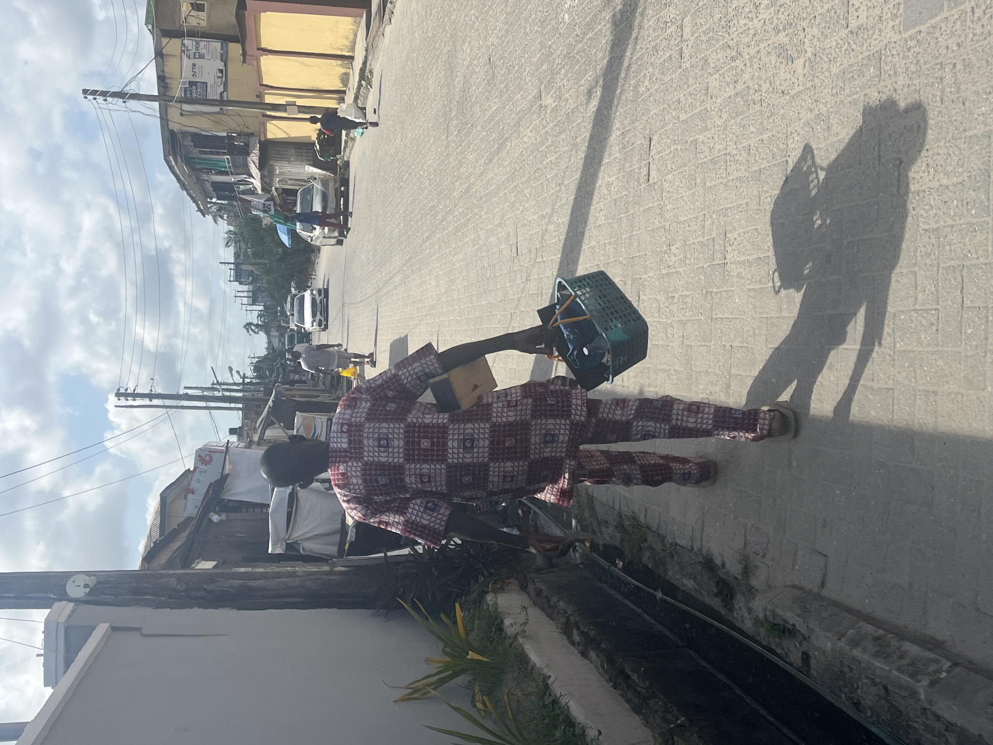 A man walks down a sunlit street in Ado, Nigeria. He wears a patterned long-sleeved shirt and pants, and sandals. He carries a green plastic basket. The street is paved with light-colored bricks. Buildings line the street, with utility poles visible. Cars are parked along the street in the distance. The man's shadow stretches across the pavement. A drainage ditch runs along the edge of the street.