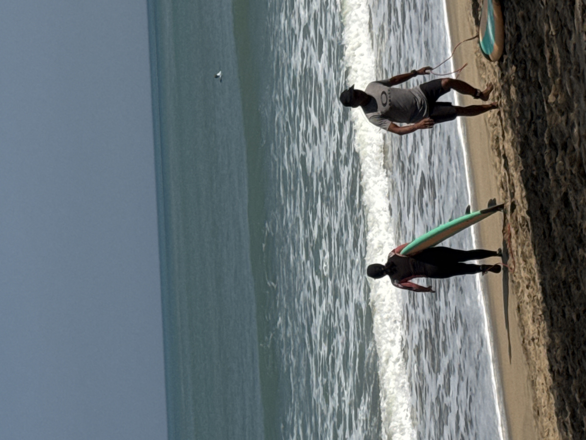 At the shore of Kuta Beach in Indonesia, two individuals, one female in a black wetsuit and the other male in a grey t-shirt and black shorts, walk with surfboards. The female, with her back to the camera, carries a green and yellow surfboard. The male, facing the camera, holds a leash attached to a surfboard lying on the sand. Gentle waves lap the shore under a clear, blue sky. A bird is visible in flight over the ocean.