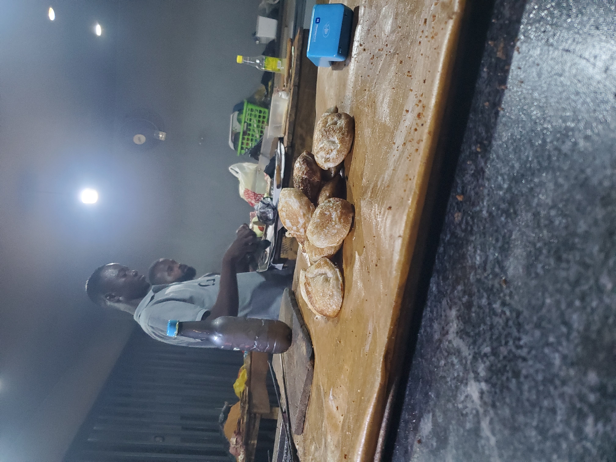 This image captures a nighttime scene at a food stall or informal eatery in Akwanga, Nigeria. The primary subjects are two men, one actively engaged in food preparation, and various food items and equipment.
The main focus is a wooden counter or cutting board on the right, laden with several cooked, golden-brown bread rolls or buns, some of which appear to be sliced open. A large, dark-handled cleaver or spatula rests on this surface near the food. In the upper right corner of the counter, a bright blue payment terminal is visible, featuring the text "PocketMoni" on its side, indicating electronic payment capabilities. Behind the counter, a yellow-capped bottle and a green plastic basket are partially seen.
On the left, two men are present. The man in the foreground, wearing a light-colored shirt and a blue wristband on his left arm, stands behind the counter. He is holding a pair of metal tongs, suggesting he is actively preparing or serving the food. Another man's head and shoulders are dimly visible behind him. Smoke or steam rises in the air, hinting at ongoing cooking activities, possibly grilling or frying.
The setting is dimly lit by several overhead artificial lights, indicating it is night or an indoor/sheltered location. The floor or ground visible is dark and appears to be made of concrete or a similar material. The scene conveys a rustic yet functional environment, common for street food vendors or local eateries.