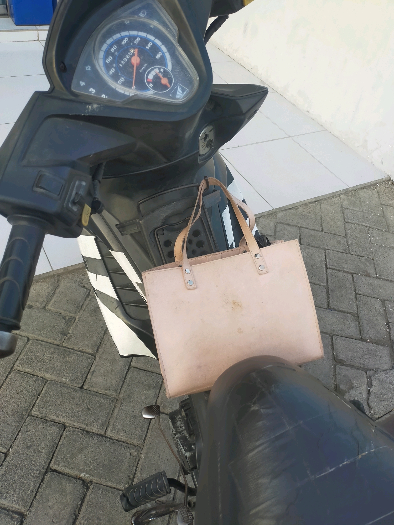 The image shows a light-colored tote bag with leather straps resting on the seat of a black motorcycle. The motorcycle's dashboard is visible, featuring a speedometer with markings up to 160 km/h and an odometer reading of 33952. The motorcycle is parked on a paved ground made of interlocking concrete blocks, and a portion of a white wall and a blue object are in the background. The scene appears to be outdoors, possibly in a public area like the vicinity of Puskesmas Keboan, Indonesia, given the tiled ground and wall which are common features in such locations. There are no people visible in the image.