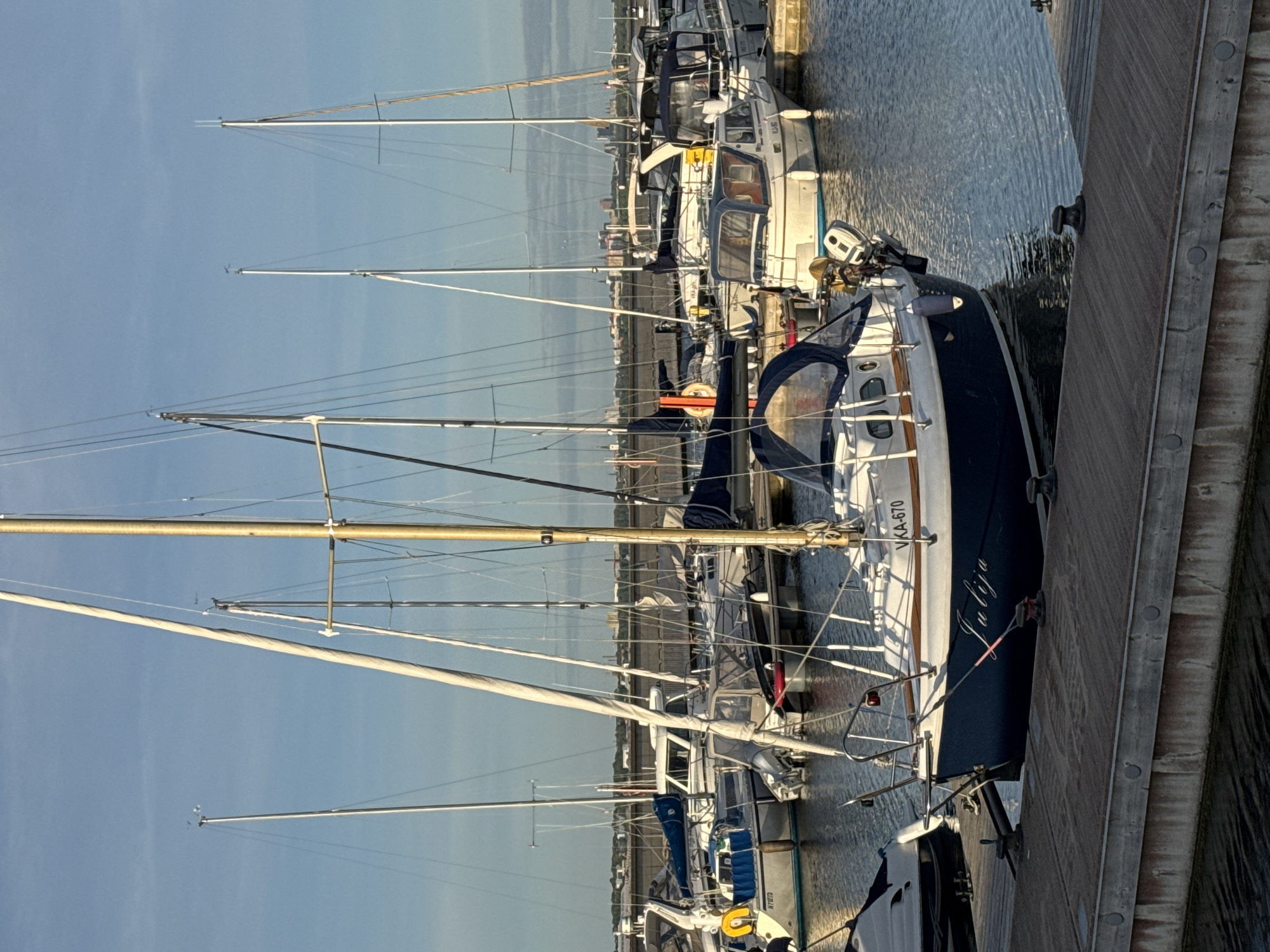 The image shows a marina in Tallinn, Estonia, with several sailboats docked. The foreground is occupied by a wooden pier with cleats. In the middle ground, a blue and white sailboat named "Julija" is prominently featured, with its registration number VKA-670 visible. Behind it, other sailboats and motorboats are moored. The background shows a distant cityscape and a clear blue sky with a few wispy clouds. The lighting suggests it is either early morning or late afternoon, with the sun casting long shadows. There are no people visible in the image. The water is calm, reflecting the sky and the boats. The overall scene depicts a peaceful harbor environment.