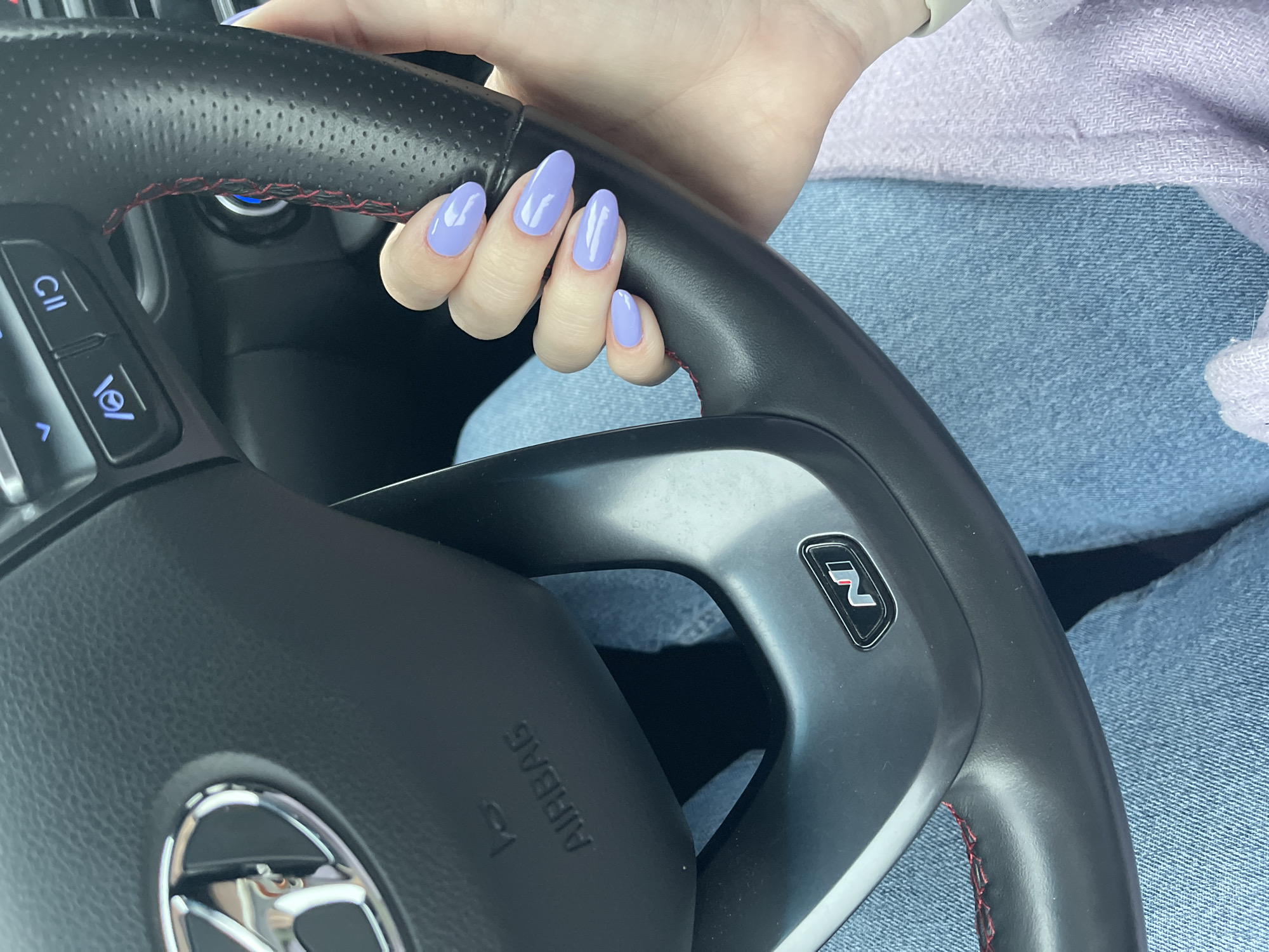 A person's hand, adorned with light purple almond-shaped nails, is resting on a black leather steering wheel with red stitching. The steering wheel has an "N" logo on it. The driver is wearing light blue denim jeans and a light purple sweater. The setting is inside of a car, but the location cannot be identified as Sofia, Bulgaria, from the provided image.