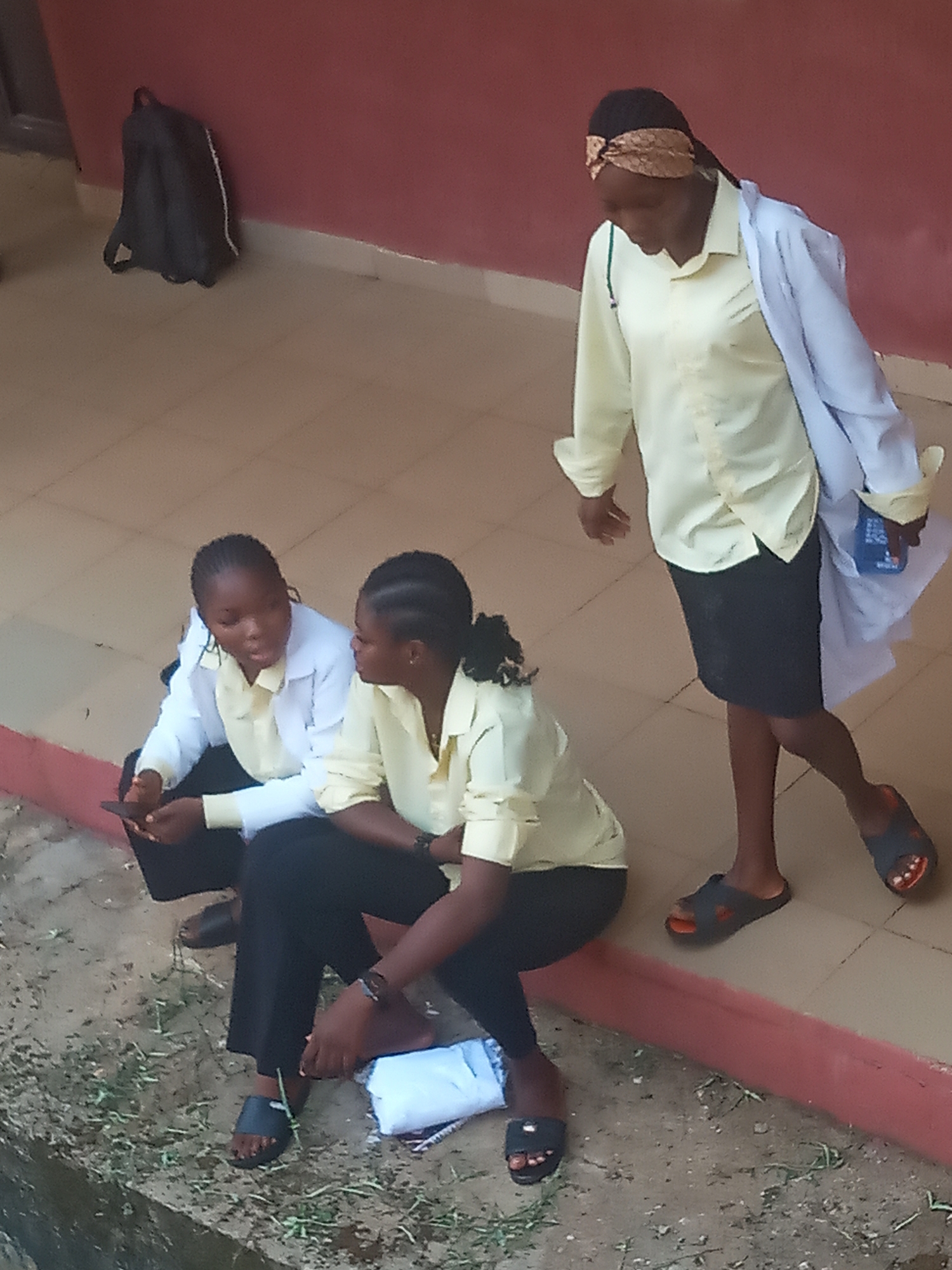 The image depicts three young women in an outdoor setting in Nkwelle, Nigeria, under natural daylight. Two of the women are seated on a low concrete ledge or step, facing each other and engaged in conversation. Both are dressed in light yellow shirts and dark bottoms, with one also wearing a white lab coat. The woman on the left is holding a mobile phone. The third woman stands to the right, wearing a light yellow shirt, a dark skirt, and a white lab coat draped over her left arm. She has a patterned headscarf and is looking downwards, holding a blue object with illegible white text. All three women are wearing sandals or slippers. In the background, a dark grey backpack leans against a reddish-brown wall with a white base trim. The ground is a mix of light-colored tiles near the building and dirt with sparse vegetation in the foreground. The scene suggests a casual interaction or break, possibly within a school or university environment.