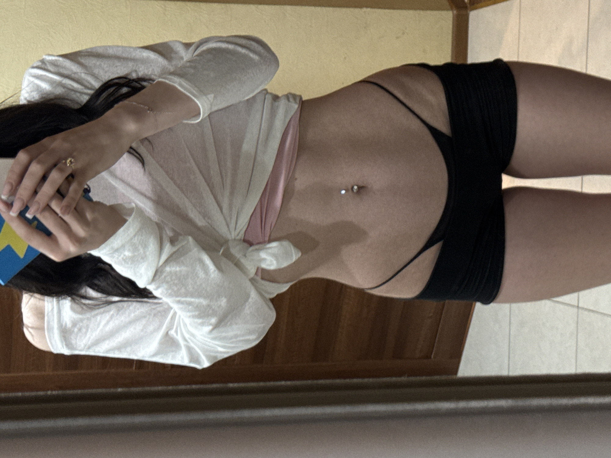 The image shows a person's torso in a room. The person is wearing an off-the-shoulder white long-sleeved shirt tied at the waist, revealing a pink undergarment. They are also wearing black shorts and a black thong. The person has a navel piercing. The background consists of a wooden door on the left and a yellow wall on the right. The floor is tiled. A mirror is visible on the left edge of the image, reflecting the scene. The image does not provide enough information to determine the location in the city of Khimki, Russia.