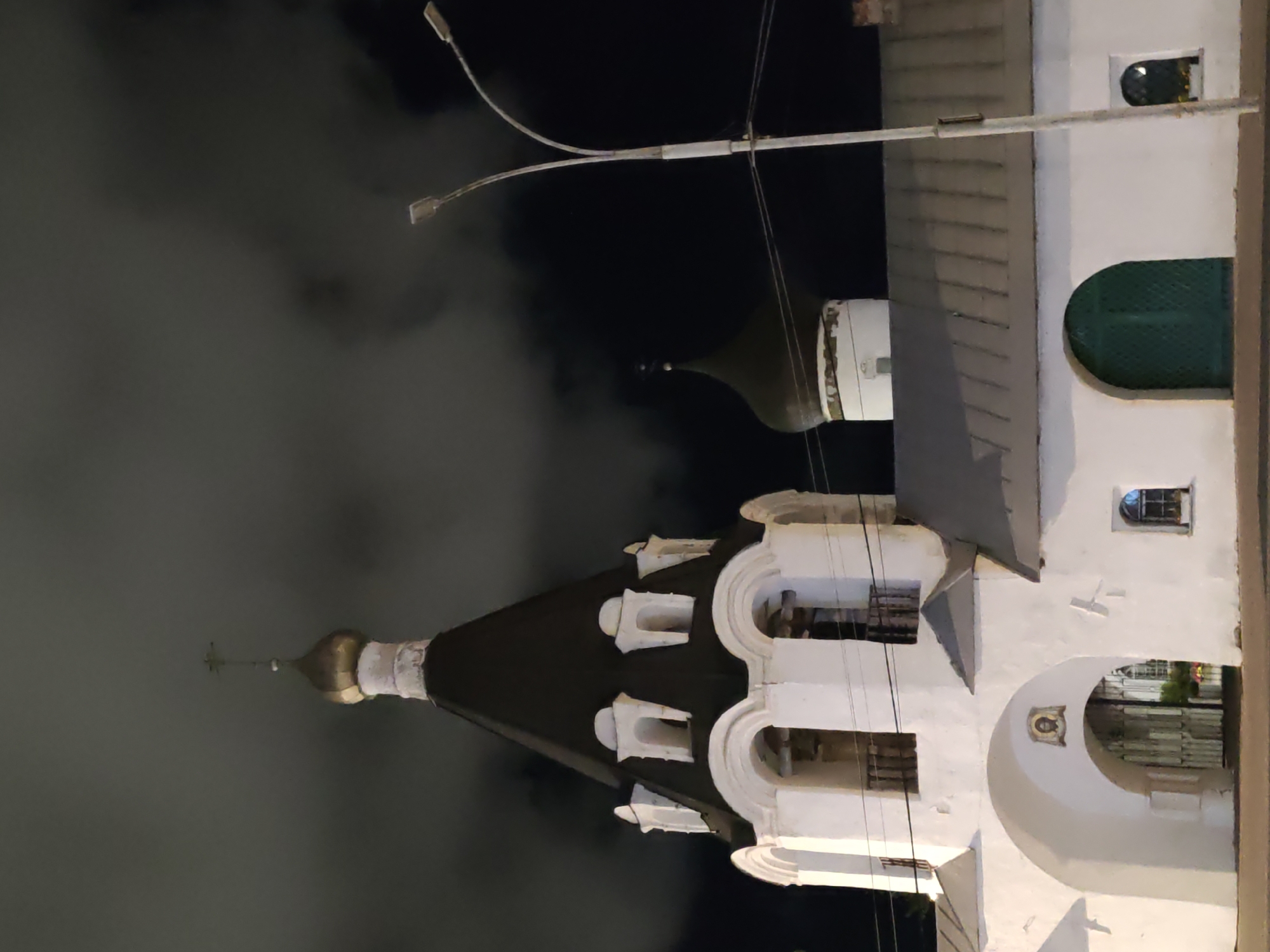 The image captures a night view of the Pskov Kremlin in Pskov, Russia, focusing on a white gate tower and a section of the surrounding wall. The gate tower features a distinctive dark conical roof topped with a gold dome and a cross. Below the dome, the tower has white arched openings with visible dark interiors, suggesting it might be a bell tower or part of the church structure. The lower section of the gate tower is a white arched entryway with a framed religious icon above it. To the right of the gate tower, a portion of the white wall of the Kremlin is visible, with a grey, sloped metal roof and a dark green arched door. Another small arched window with a flower box is also present on this section of the wall. In the background, a dark, domed structure, likely another church within the Kremlin, is partially visible. A modern street lamp with two lights stands to the right of the gate tower, its pole and arms illuminated. The sky is dark and cloudy, with the overall lighting suggesting it is nighttime, with artificial light sources illuminating the architectural elements.