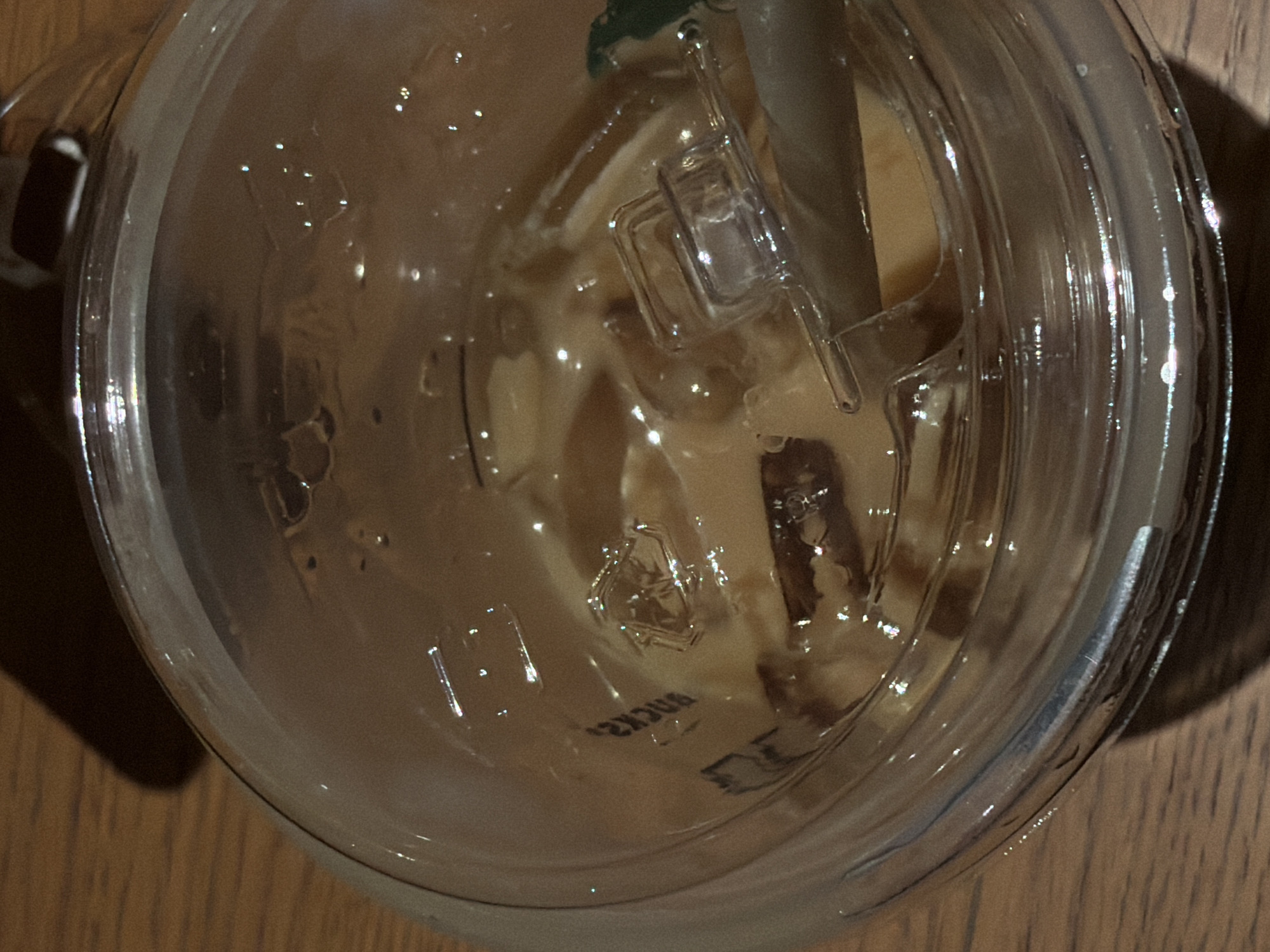 A close-up, overhead view of a clear plastic cup filled with a light brown, iced beverage, likely coffee or tea, sits on a wooden surface. The cup has a ribbed texture on its outer surface. Inside the cup, ice cubes and condensation are visible. A green straw with a clear plastic lid is partially submerged in the drink. The lid has a small lever mechanism for opening and closing the straw hole. There appears to be some text embossed on the bottom of the cup, which is partially obscured by the liquid and ice, but the letters "BOCK" are discernible, possibly part of a brand name. The lighting suggests an indoor setting, with reflections and highlights glinting off the cup and liquid. The wooden table provides a warm, natural backdrop. The overall impression is of a refreshing beverage ready for consumption. The scene is static, capturing a moment in time. There are no people visible in the image. The provided location context of "Đà Nẵng, Vietnam" does not offer specific visual cues within the image itself, as the scene is generic and could be anywhere.