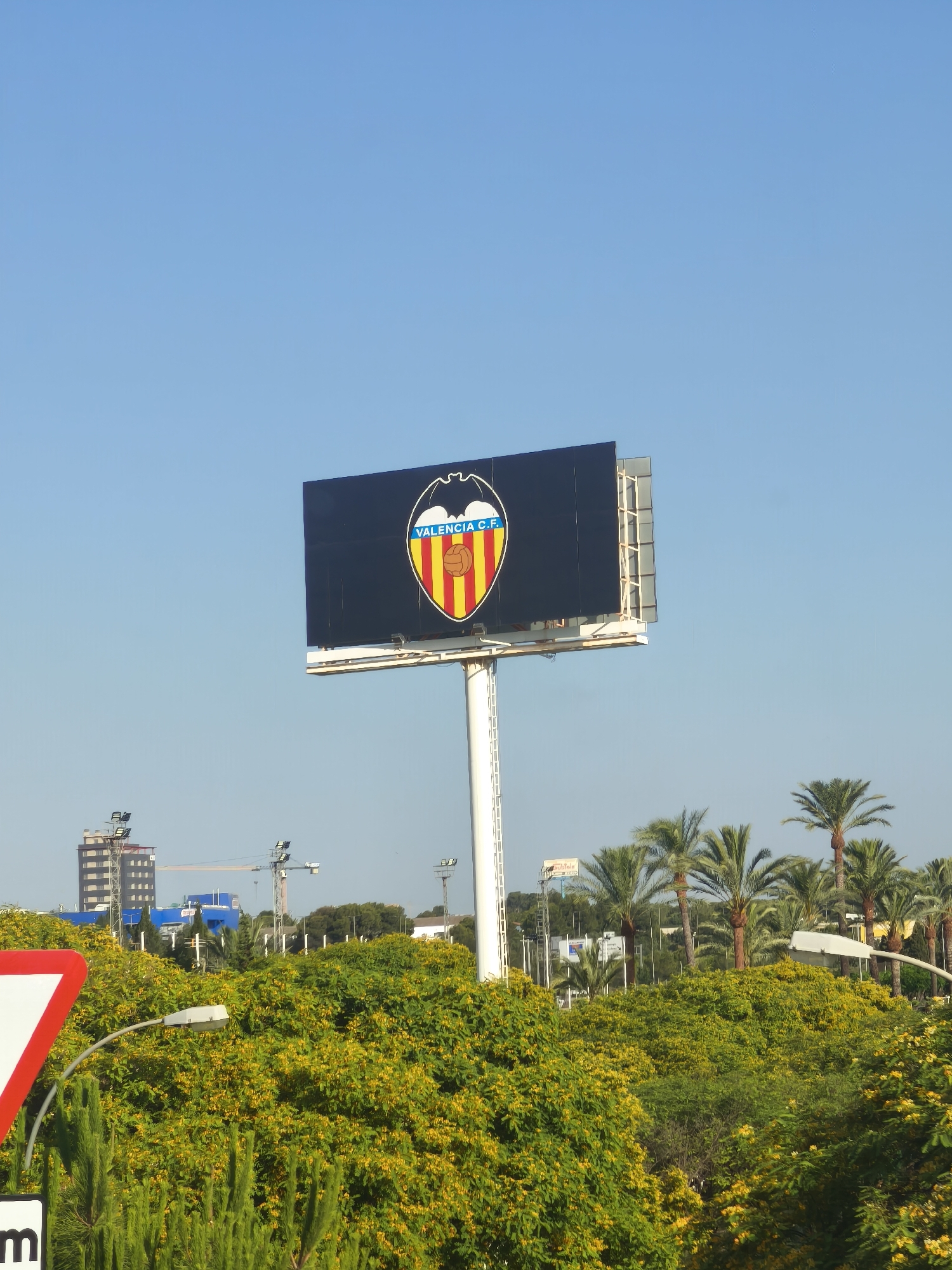 A wide shot of a clear blue sky above a billboard displaying the Valencia C.F. crest, supported by a tall white pole. The billboard is situated in a park-like setting with lush green trees and vegetation, some bearing yellow flowers, in the foreground and mid-ground. In the background, a tall, modern building with a dark facade stands to the left, alongside construction cranes and other smaller buildings. Palm trees are visible to the right of the billboard. A red and white triangular traffic sign is partially visible in the bottom left corner, along with a black sign with the letter "m". The scene appears to be outdoors during the day, likely in a city environment, possibly Paterna, Spain, given the presence of the Valencia C.F. crest.