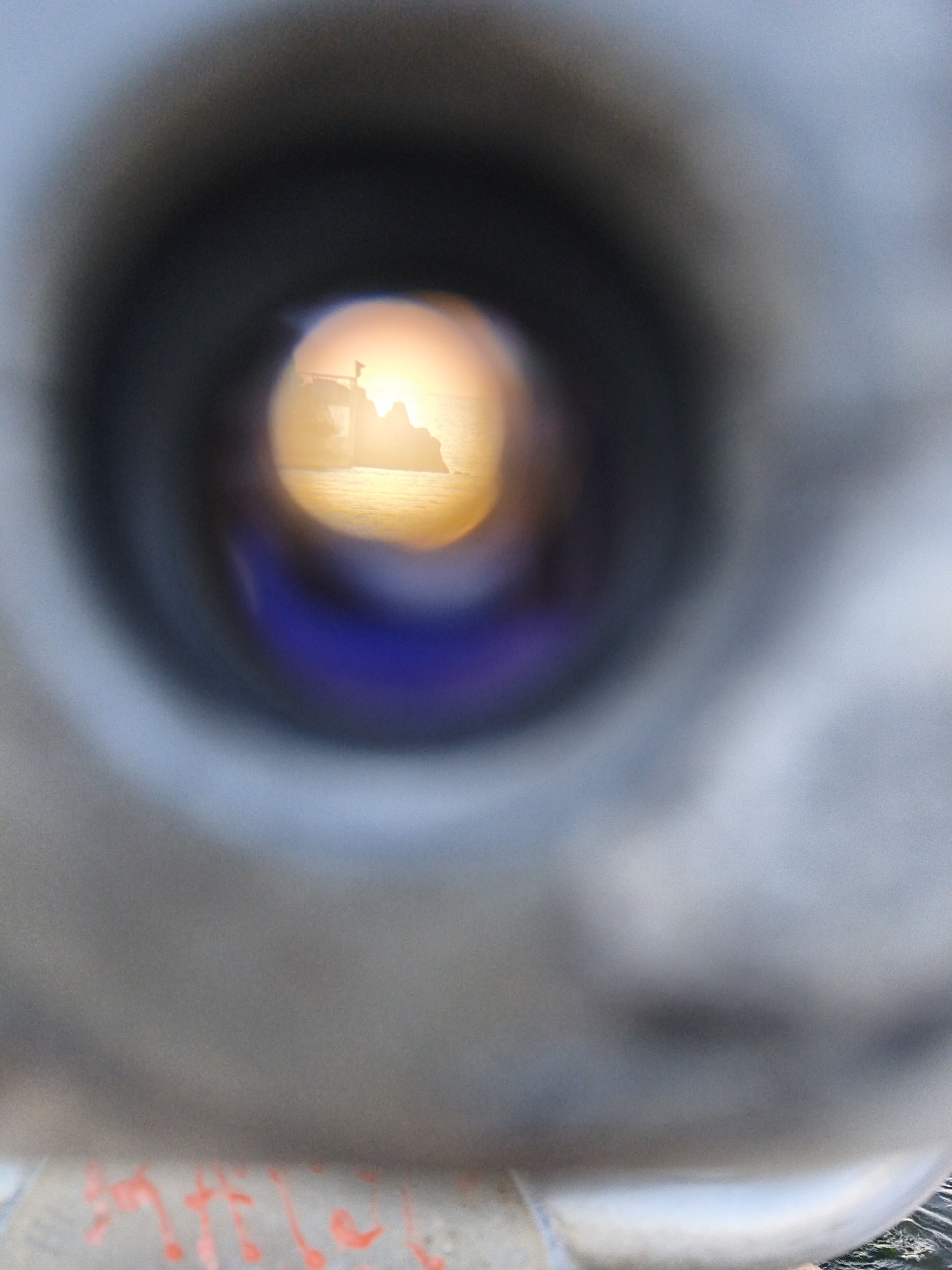 The image is a close-up shot looking through a viewing scope, revealing a distant scene. The foreground is dominated by the blurred, gray, cylindrical structure of the scope itself, with the aperture at the center showing the captured image. The aperture is circular and out of focus, framing a brightly lit horizon.
The scene within the scope appears to be a coastal landscape. A silhouette of jagged rocks or cliffs is visible against a bright, golden-orange sunset or sunrise. The sun itself is a radiant orb, casting a warm glow over the water and the rocky formations. There's a suggestion of a ship or boat in the distance, its outline faintly visible against the bright sky. The water reflects the warm light, creating a shimmering effect. Below the bright horizon, a deep blue color is visible, possibly representing the sea or sky.
The bottom portion of the original image shows a blurry surface with bright red paint streaks, possibly graffiti or markings on the exterior of the viewing scope or its base. In the far lower right corner of the original image, a glimpse of a city landscape is visible, with buildings and structures suggesting an urban environment.
Given that this image was taken through a viewing scope, and considering the context of tourist attractions with viewing scopes in Tallinn, Estonia, it is highly probable that this image was captured from a viewpoint overlooking the Baltic Sea, possibly near a harbor or a waterfront promenade. Tallinn's historic Old Town and its proximity to the sea make it a common location for such viewing installations. The composition suggests a scenic spot frequented by tourists.