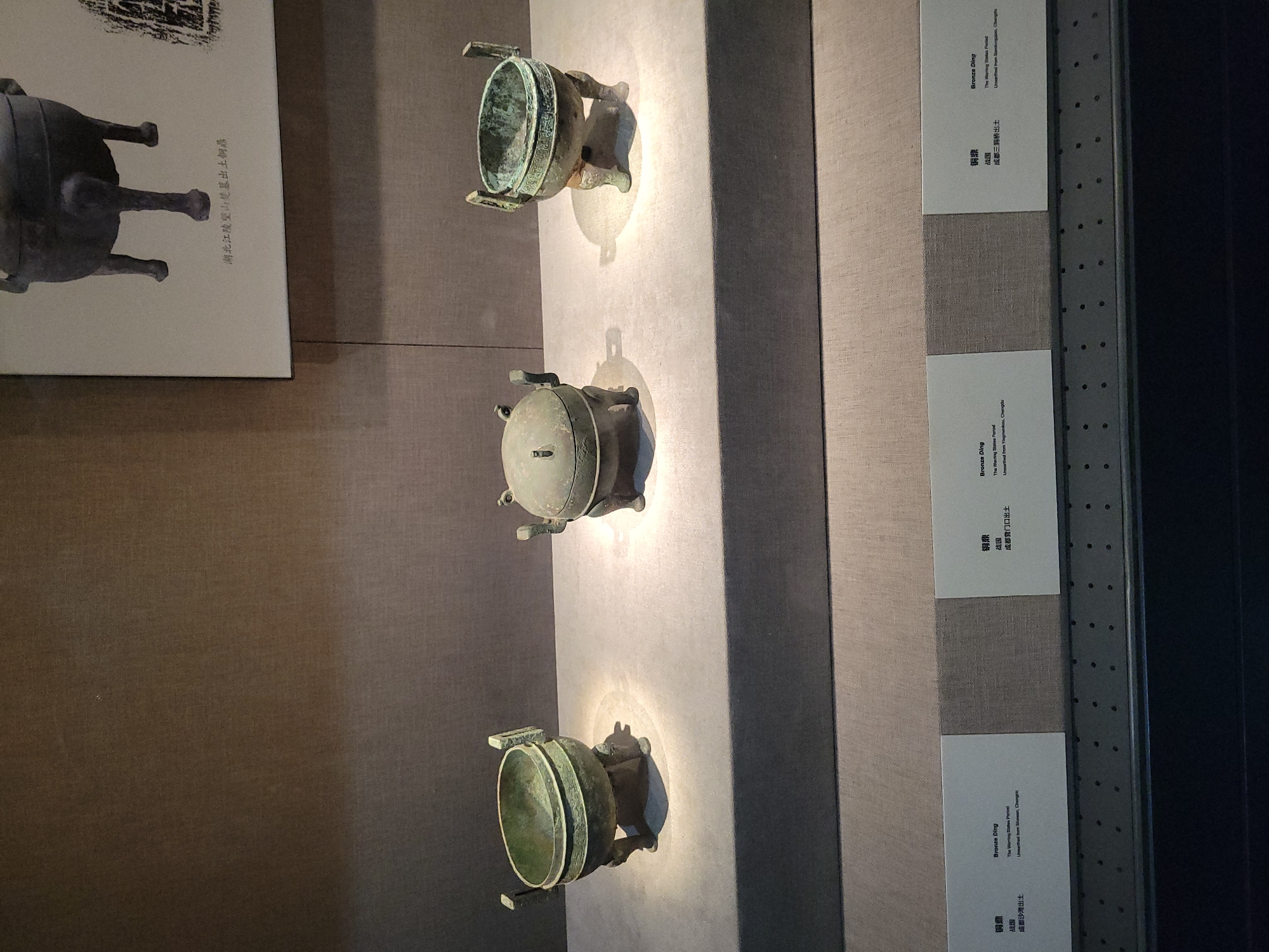The image shows three ancient bronze ding vessels displayed on a grey pedestal in a museum. The vessels are different sizes and shapes, but all have a round body and three legs. The ding in the middle is the largest and appears to have a lid. The vessels are illuminated by spotlights, casting shadows on the pedestal. In the background, a framed picture shows another bronze ding with legs. Below the pedestal are three white labels with text in Chinese and English, indicating that the vessels are "Bronze Ding" from the "Warring States Period". One label indicates it was "Unearthed from Shaoxiangkou, Chengdu". The overall environment is dimly lit, typical of a museum exhibit.